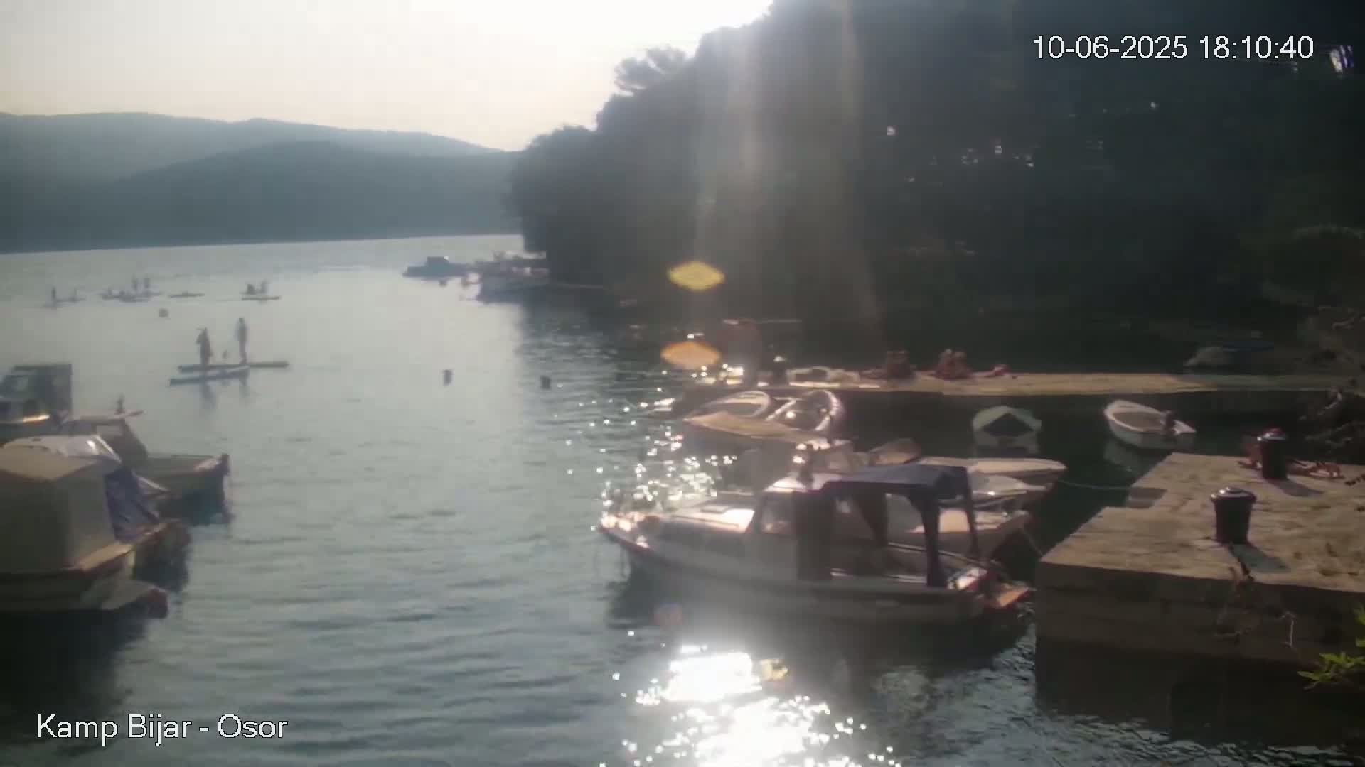 A sunny day shows several people paddleboarding in a calm bay filled with numerous small boats docked along a stone embankment.