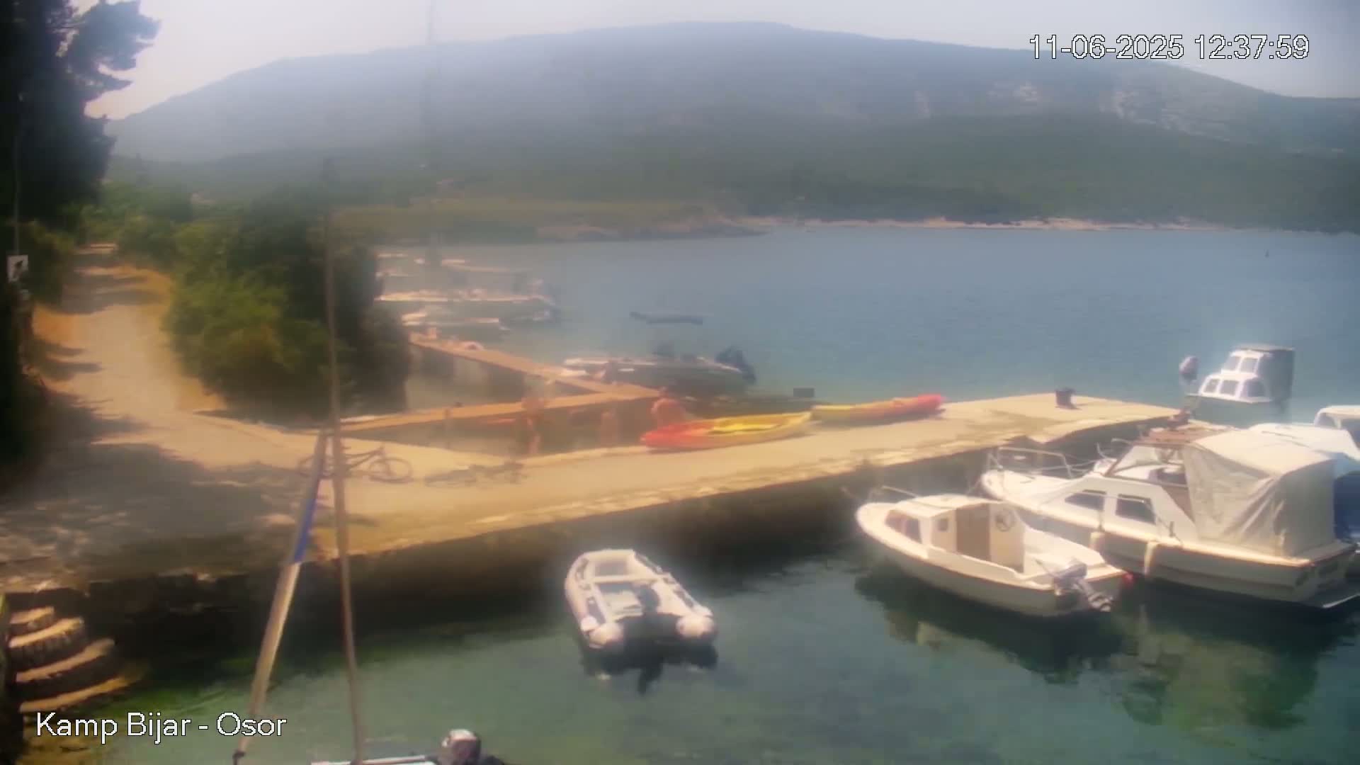 Several boats are moored at a dock near a calm bay under a hazy, sunny sky, with a path and some people visible on land.