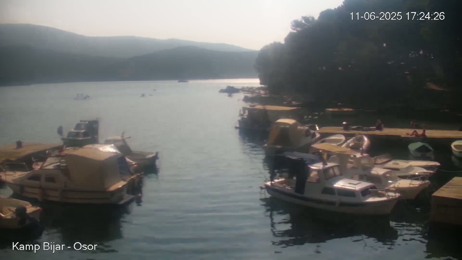 A hazy, sunny day shows numerous boats moored at a calm bay, nestled between a hillside and a wooded shore where people relax.