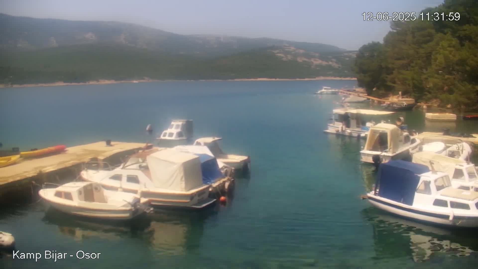 Several small motorboats are moored in a calm, clear bay, with hills and green trees visible in the background under a bright, sunny sky.