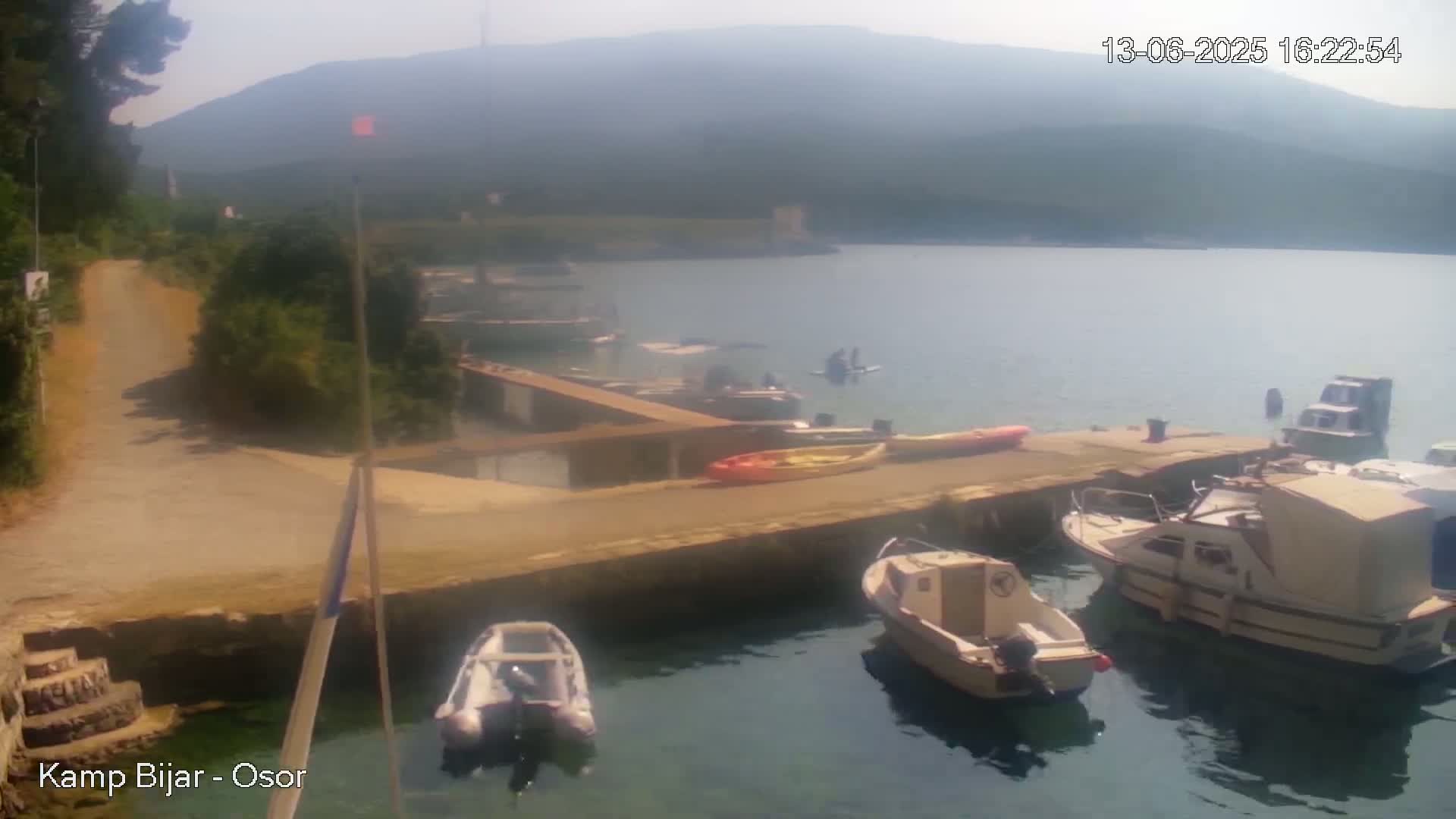 Several boats are moored at a dock on a calm body of water, with a hazy mountain visible in the background under a sunny sky.