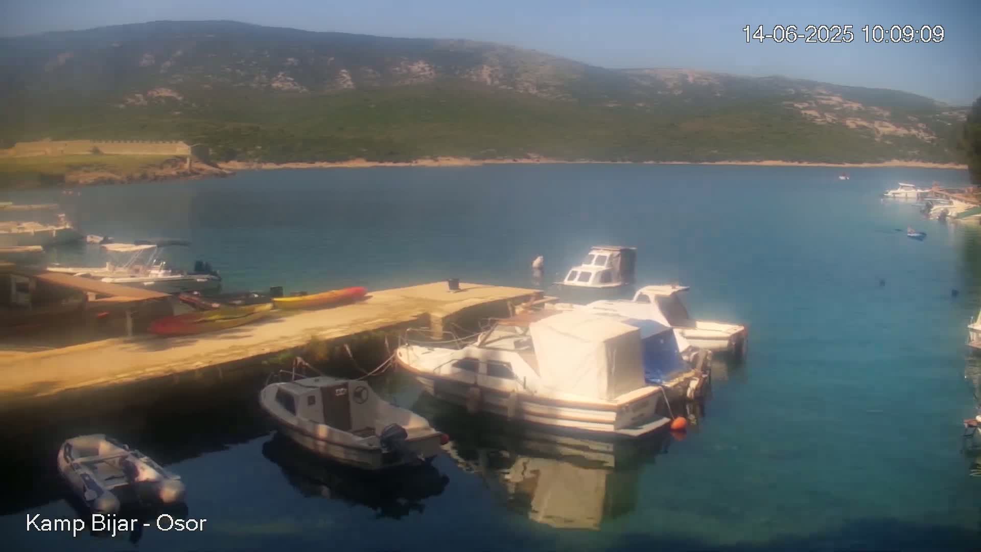 Several boats are moored at a dock in a calm, clear bay, with a mountainous, green landscape visible in the background under a sunny sky.