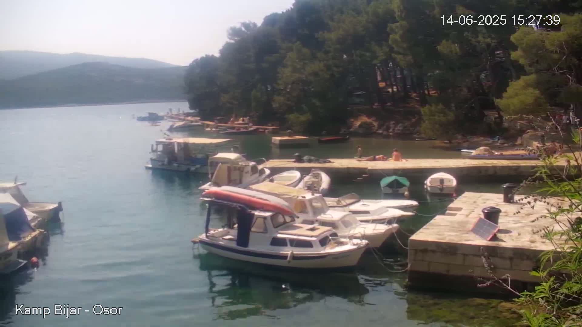 Several boats are moored at a small harbor nestled in a cove surrounded by green trees and hills under a sunny sky.