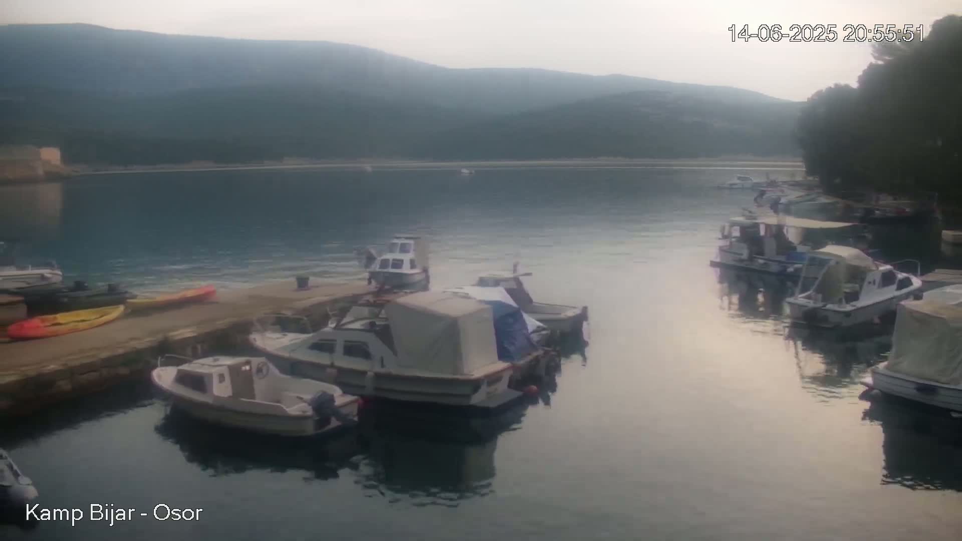 Several boats are moored at a small harbor nestled in a cove surrounded by green trees and hills under a sunny sky.