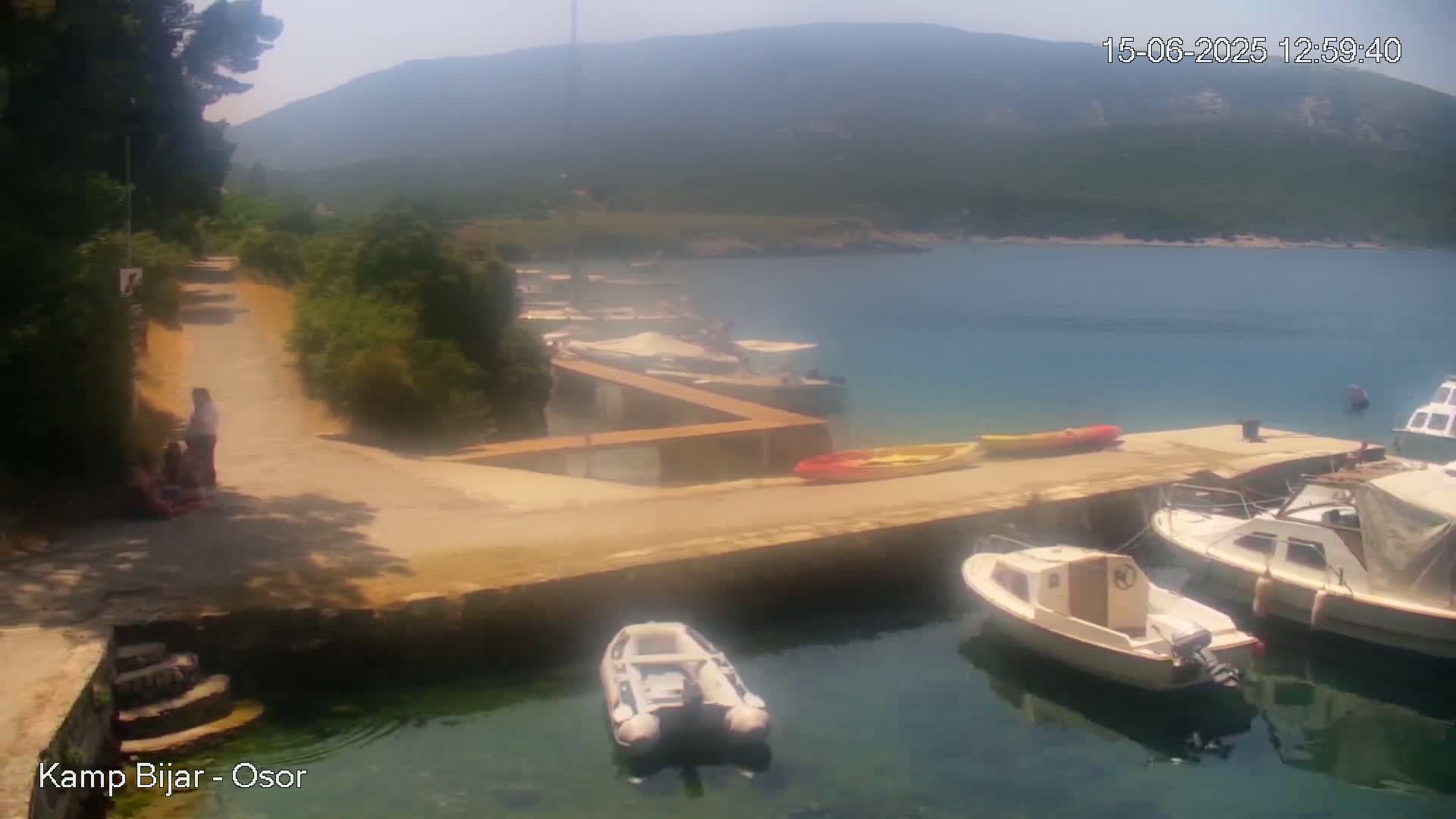 A sunny day shows a calm bay with several boats docked at a cement pier, a paved path leading from the pier to a forested area with people standing near it, and mountains in the background.