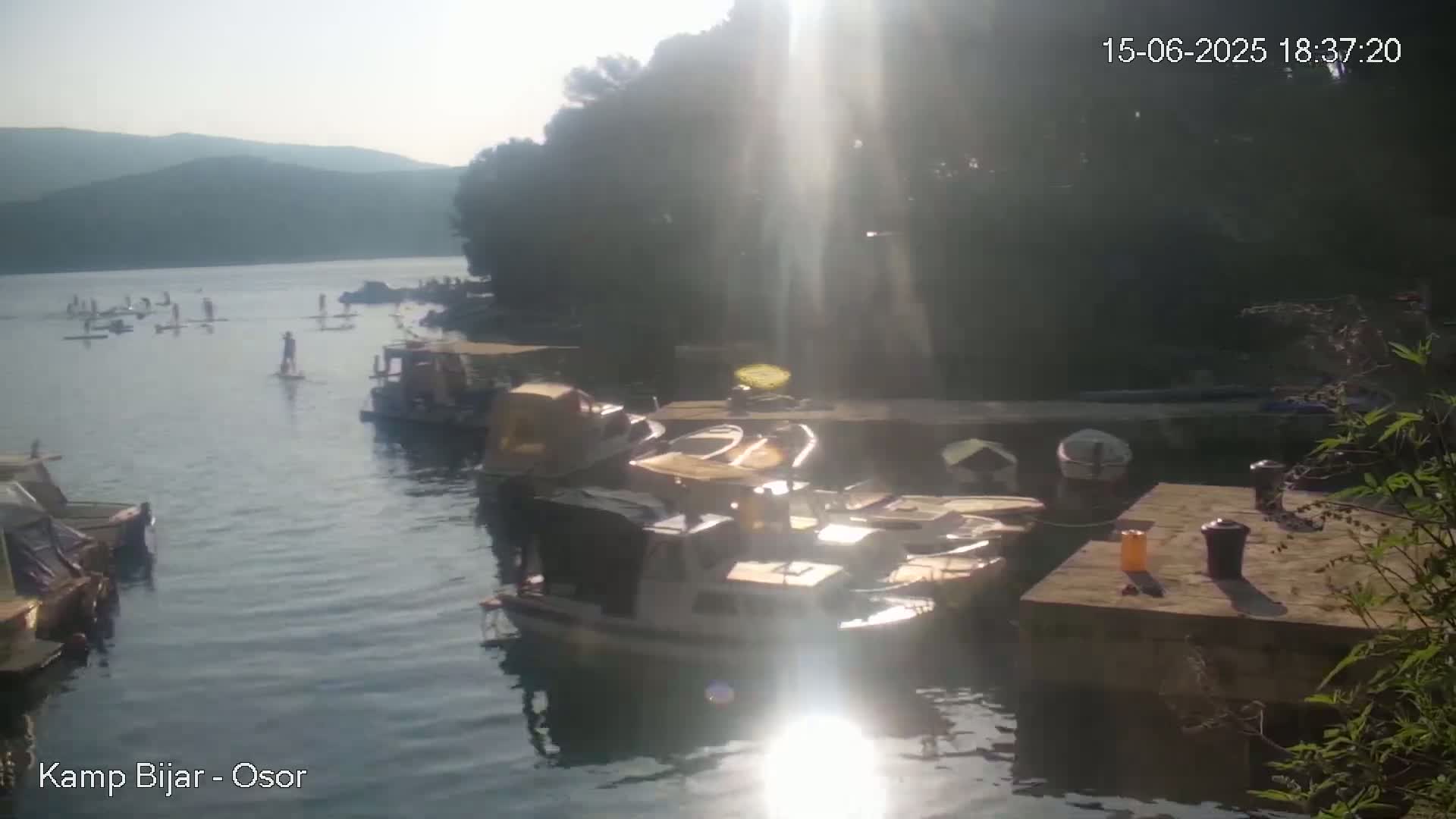 A calm body of water with paddleboarders and numerous boats docked near a shoreline under a bright, sunny sky.