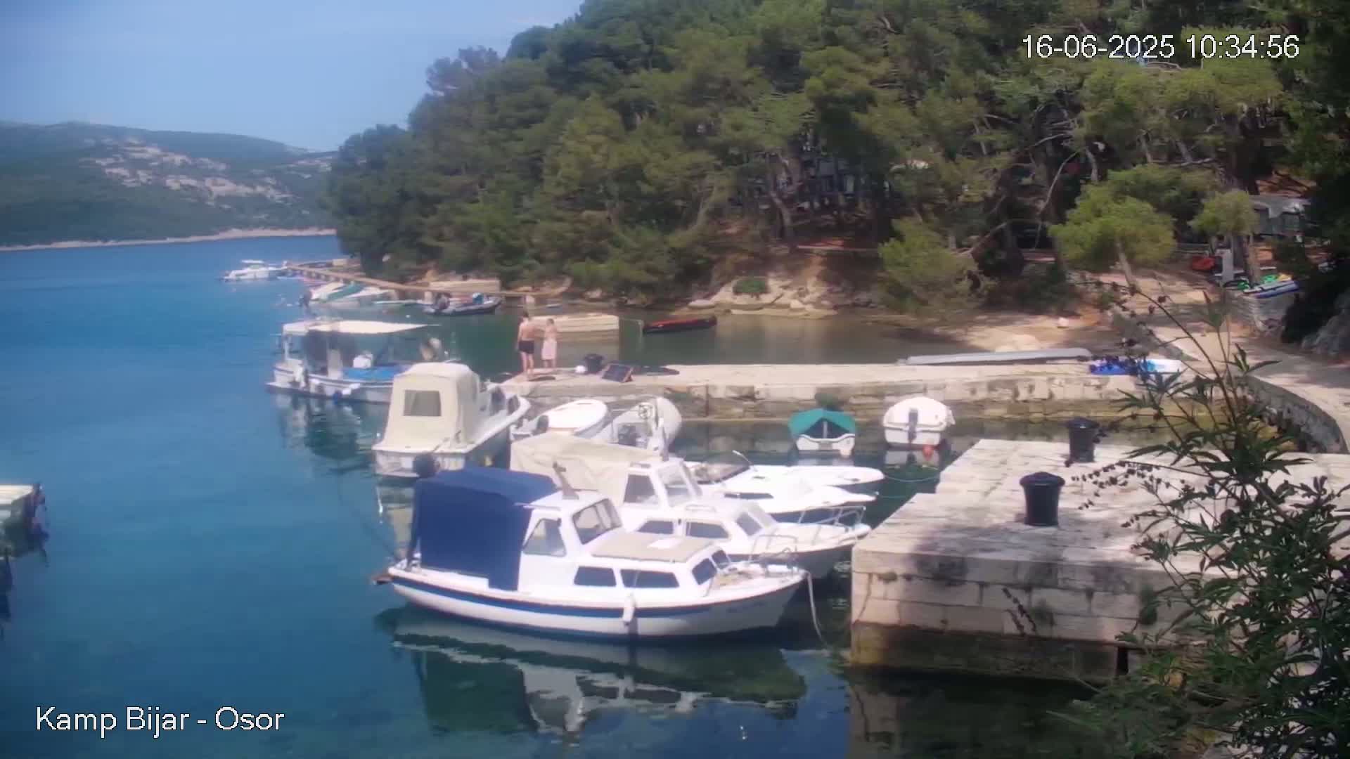 Several small motorboats are docked at a stone quay in a calm, clear bay under a sunny sky, surrounded by lush green vegetation.