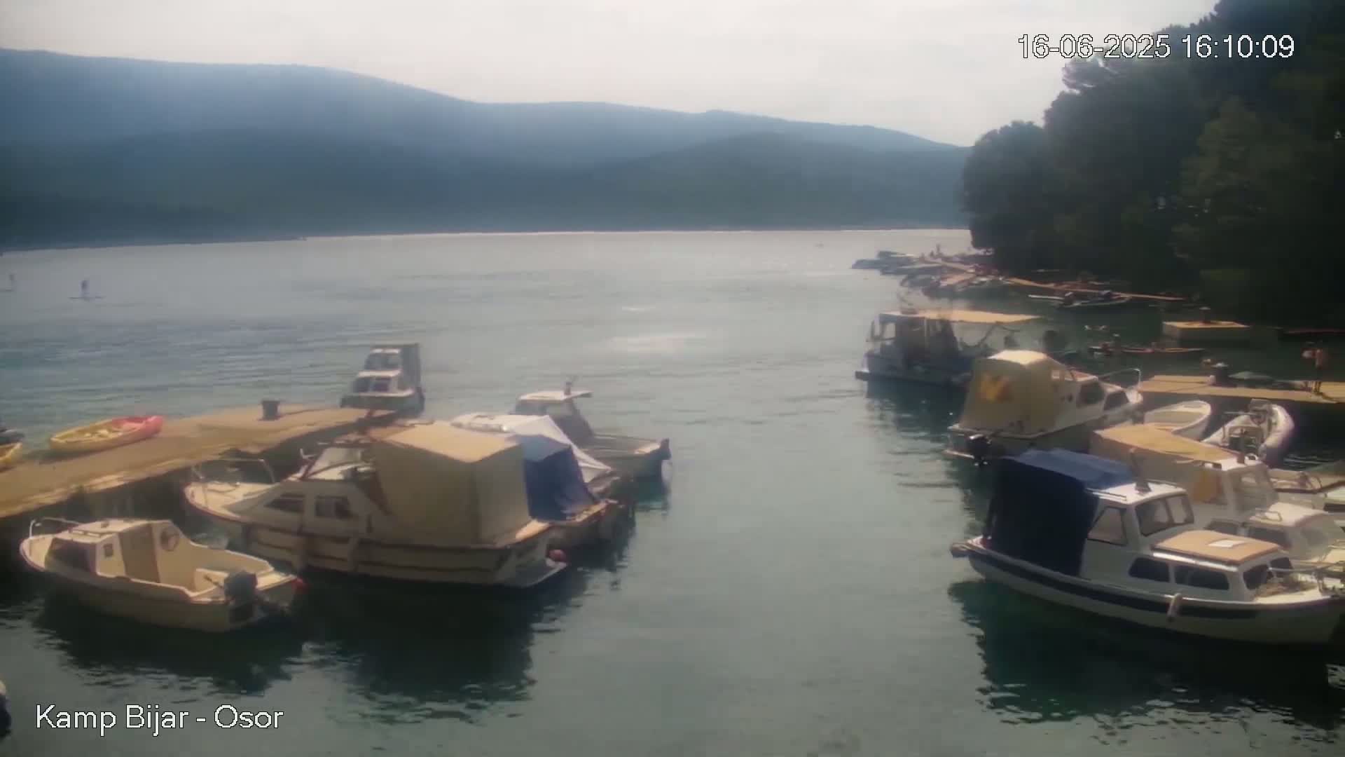 Numerous small boats are moored in calm water near a shoreline, with mountains visible in the background under a sunny sky.