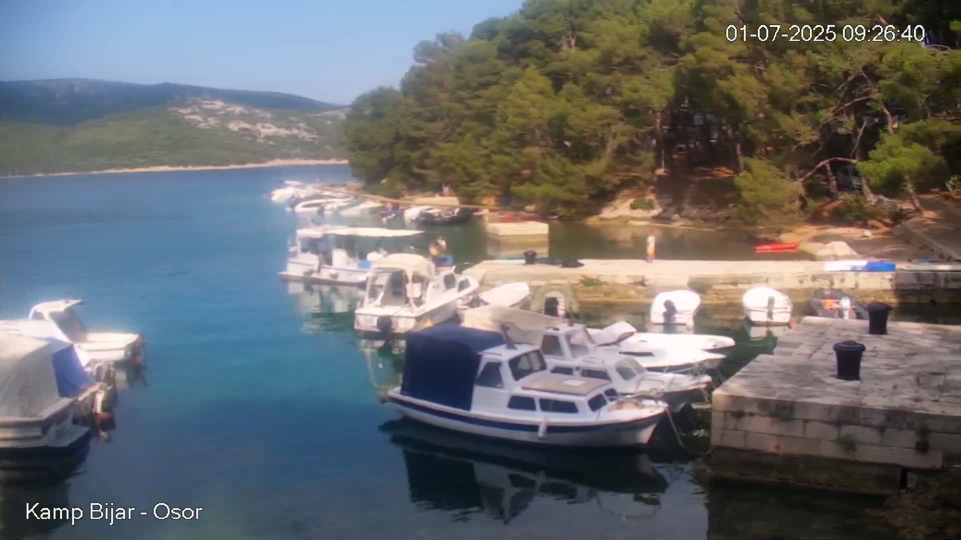 Several small motorboats are moored at a stone dock in a calm, clear bay under a sunny sky, with green hills visible in the background.