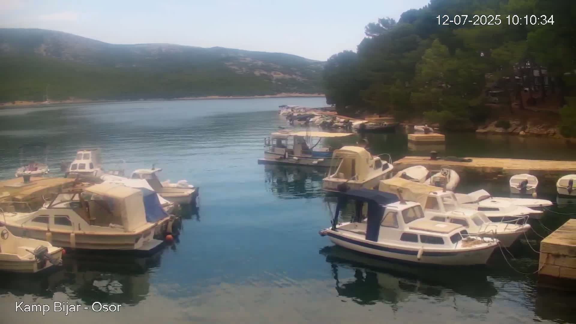 Numerous small boats are moored in a calm, clear bay on a sunny day, with verdant hills visible in the background.