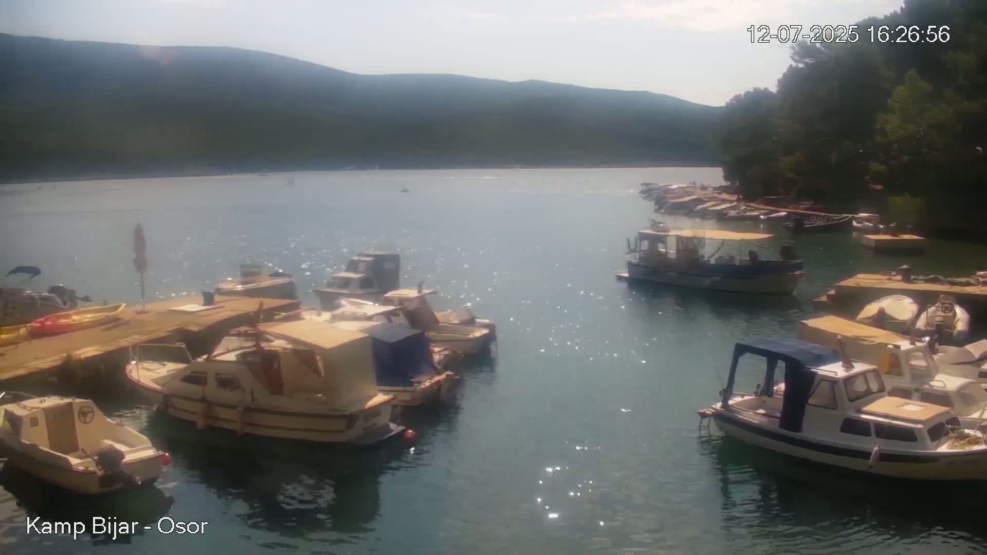 Numerous small boats are moored in a calm, clear bay on a sunny day, with verdant hills visible in the background.