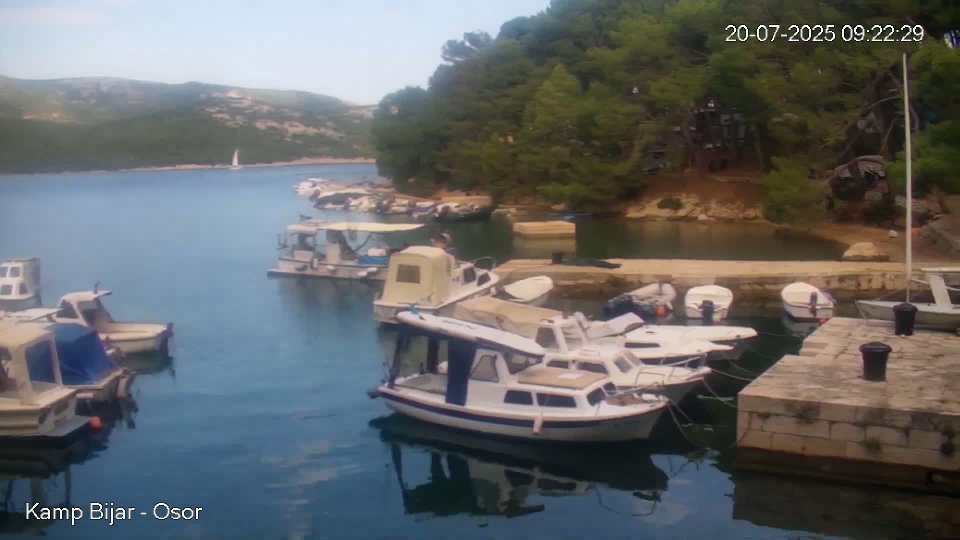 A calm bay with numerous small motorboats docked near a stone wall, nestled beside a lush green hillside under a mostly clear sky.