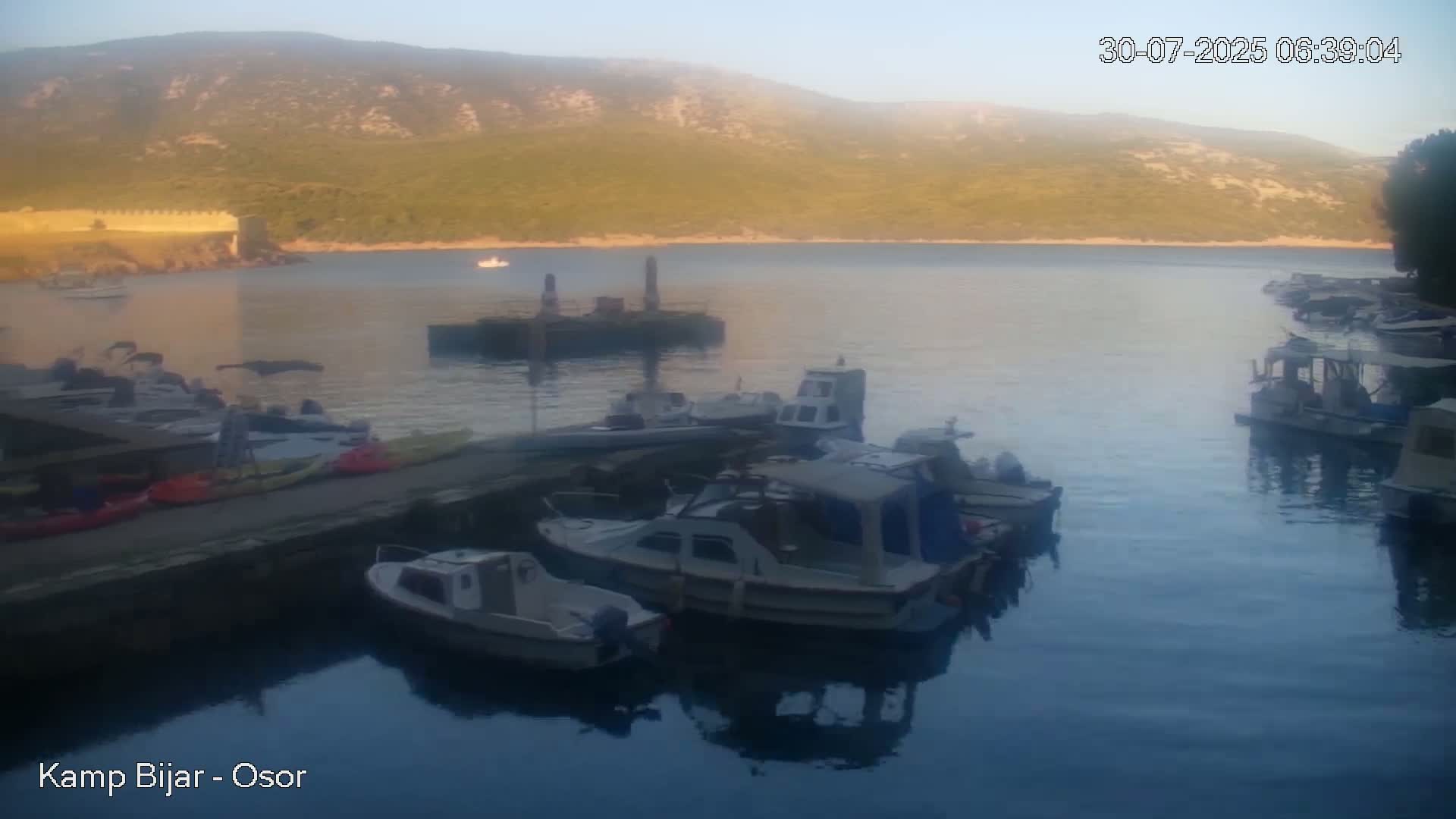 A calm bay with numerous small motorboats docked near a stone wall, nestled beside a lush green hillside under a mostly clear sky.
