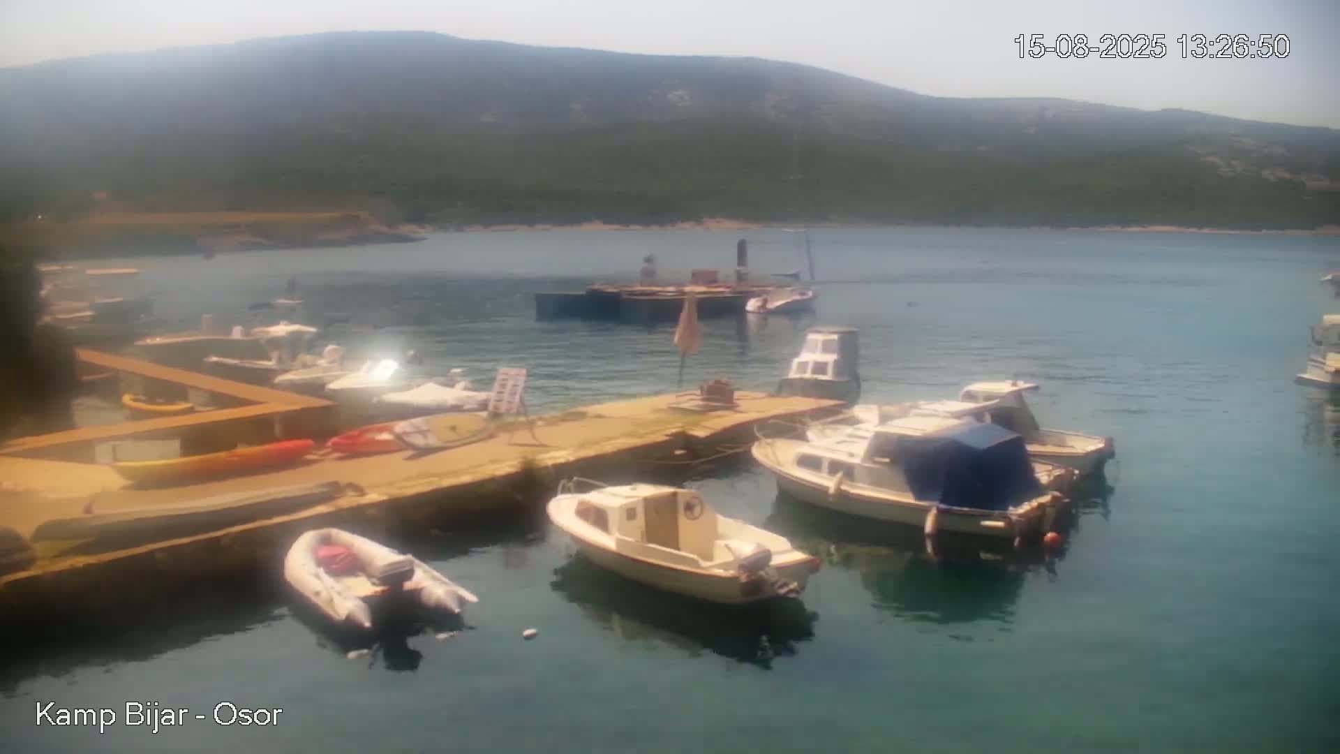 Several small motorboats are moored at a calm, sunny waterfront next to a stone dock where a few people stand near some kayaks.