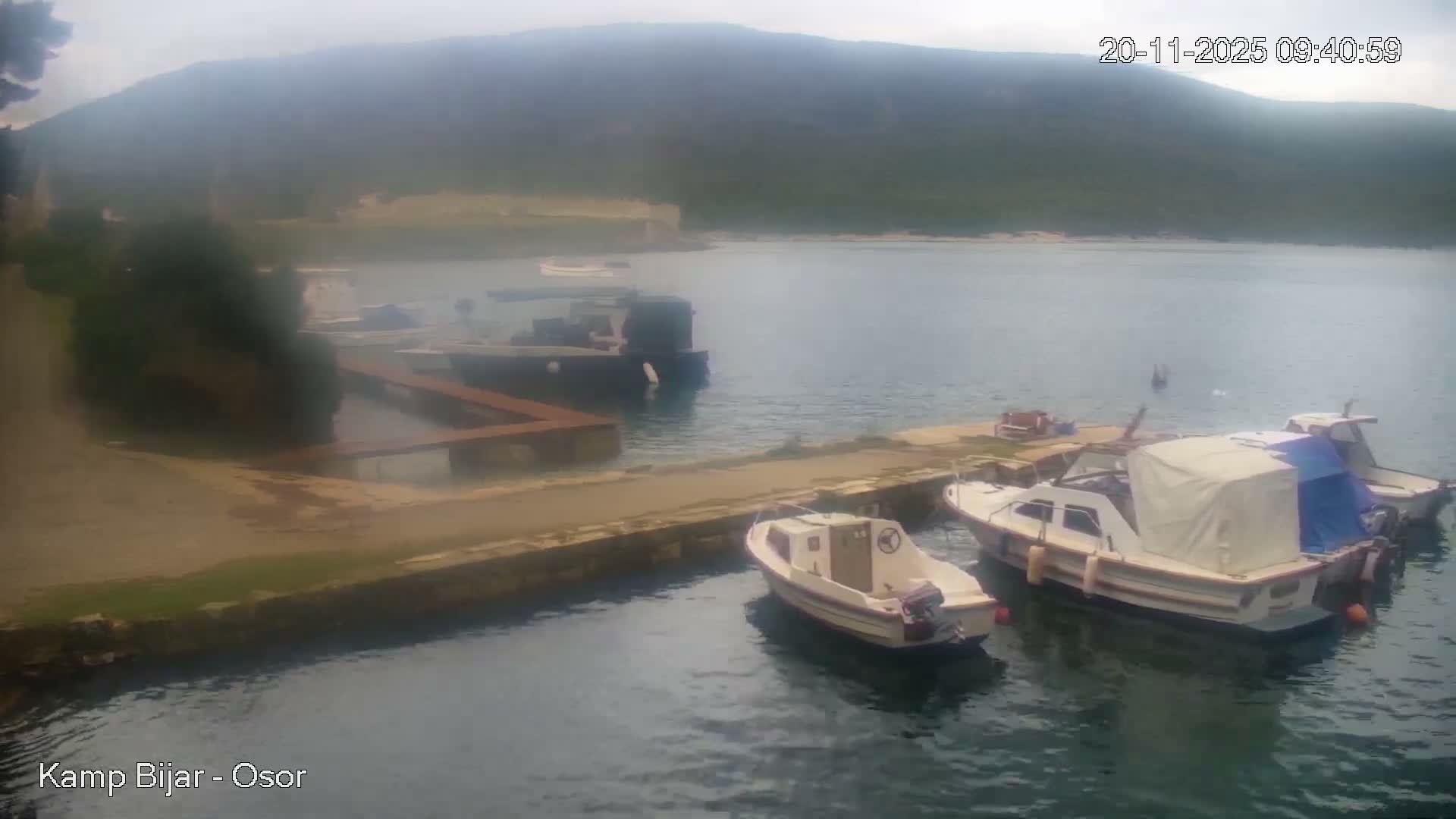 Several boats are moored along a stone quay in a calm bay, with distant hazy mountains visible under an overcast sky.