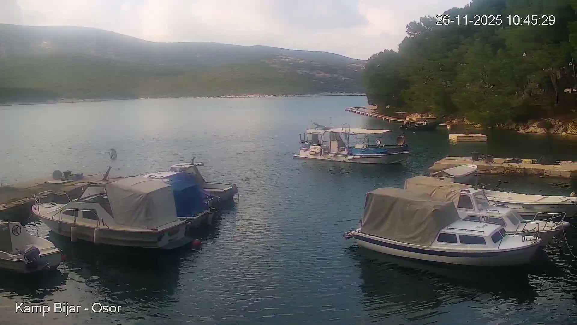 A serene bay is pictured on an overcast day, featuring several boats docked or moored, including one larger boat in the calm water, against a backdrop of distant hazy hills and a forested shoreline with multiple jetties.