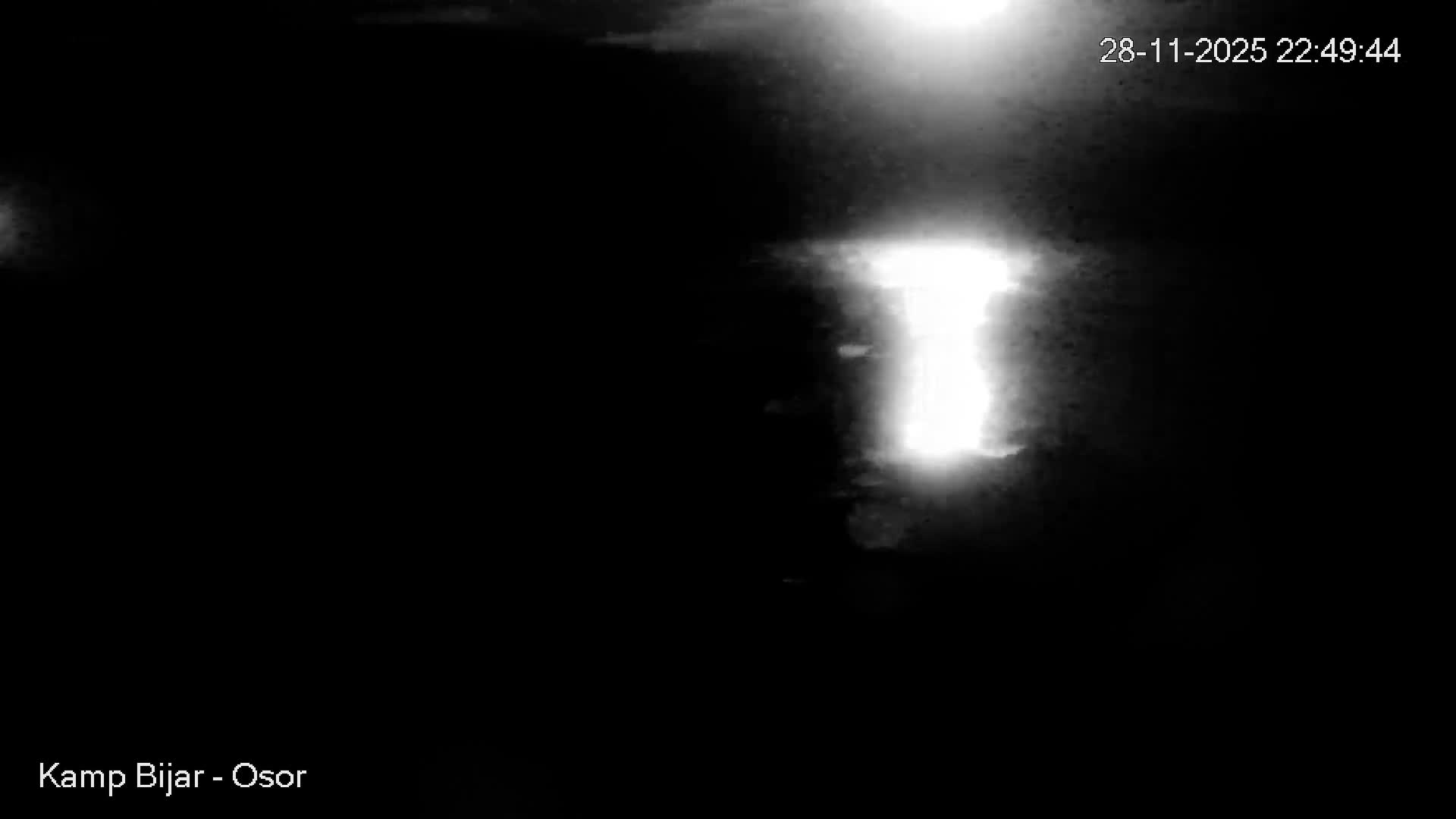 A very dark and blurry monochromatic image, taken outdoors at night, depicts a bright, diffuse light source reflecting as an elongated vertical streak on a dark, rippling water surface, possibly under misty conditions.