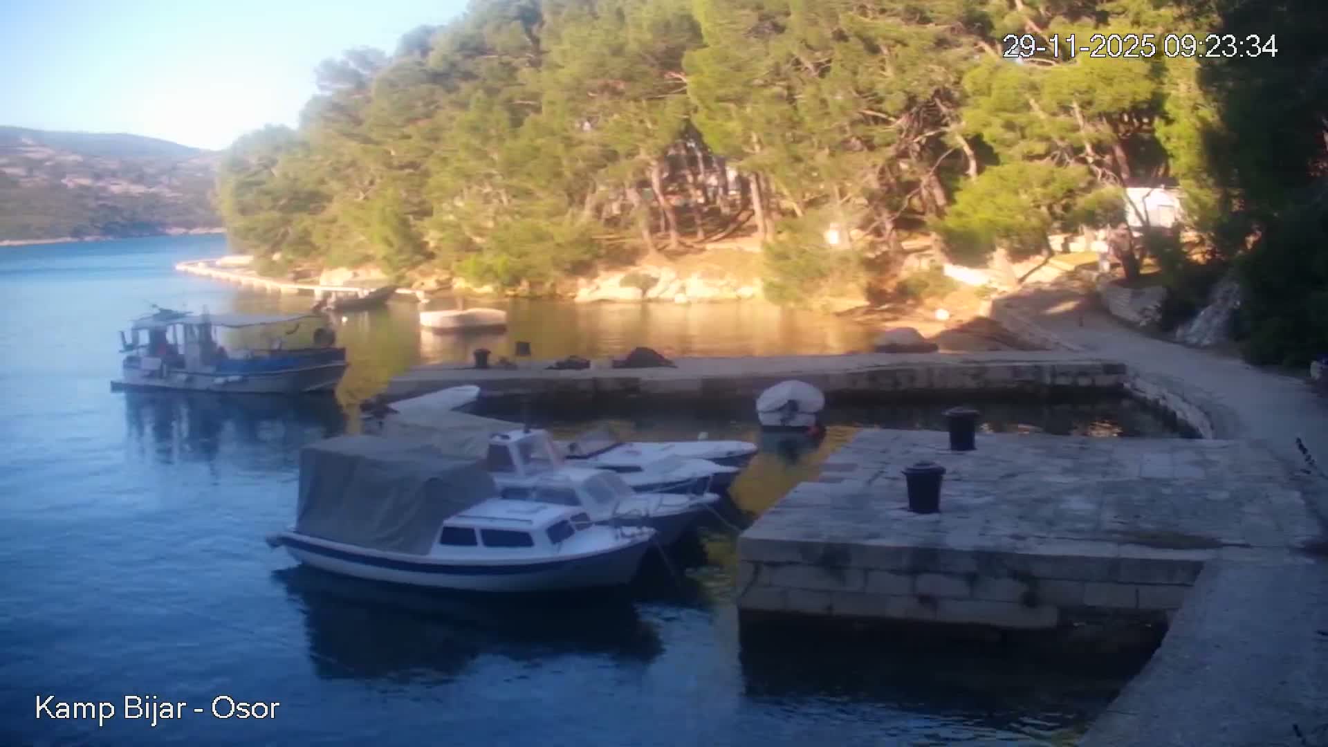 A serene coastal scene shows several boats docked at a stone pier and in the water, surrounded by clear calm water and pine-covered hills under bright, sunny skies.