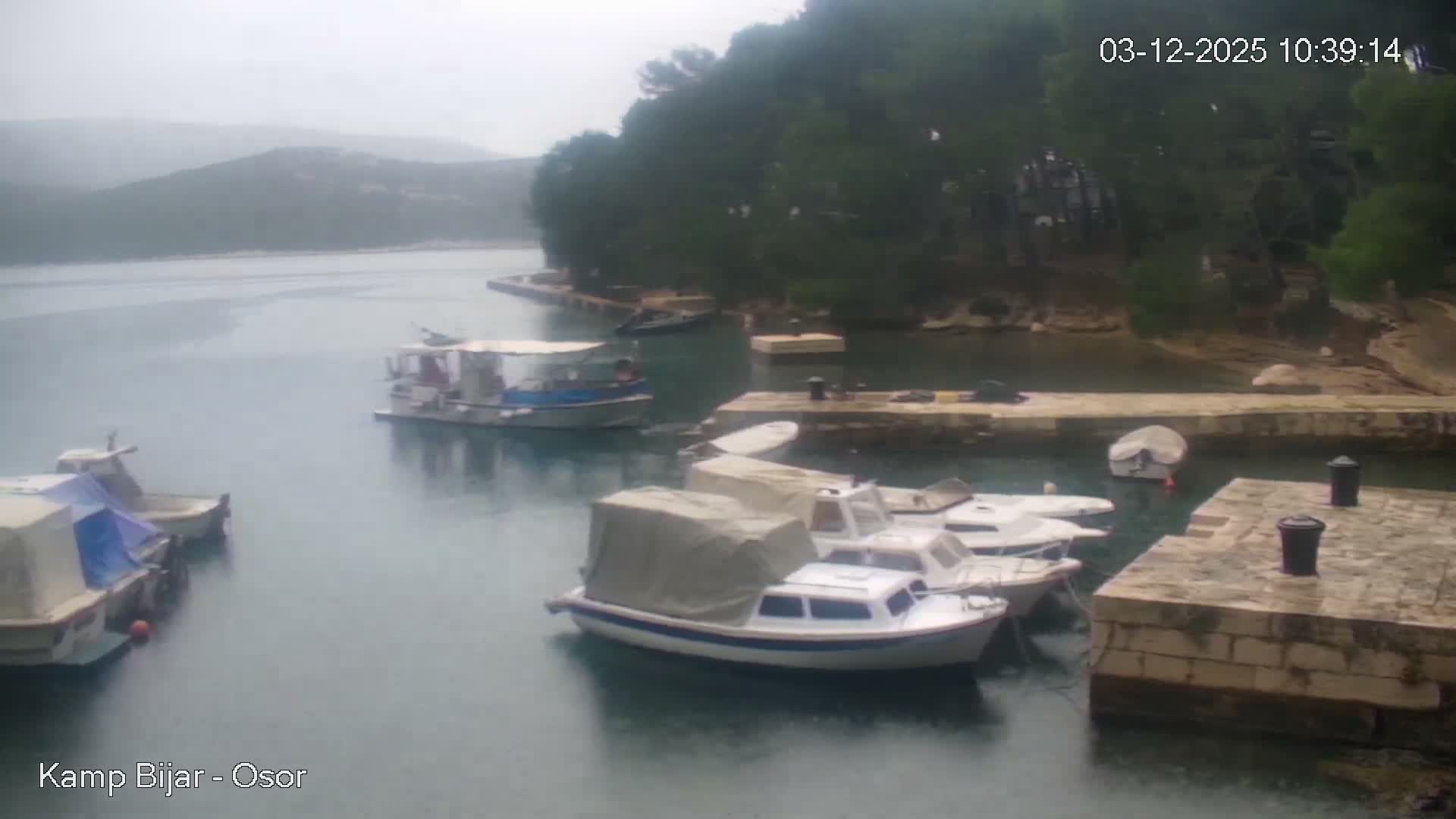Several boats are moored in a tranquil bay with stone docks and lush hills, under an overcast and rainy sky.
