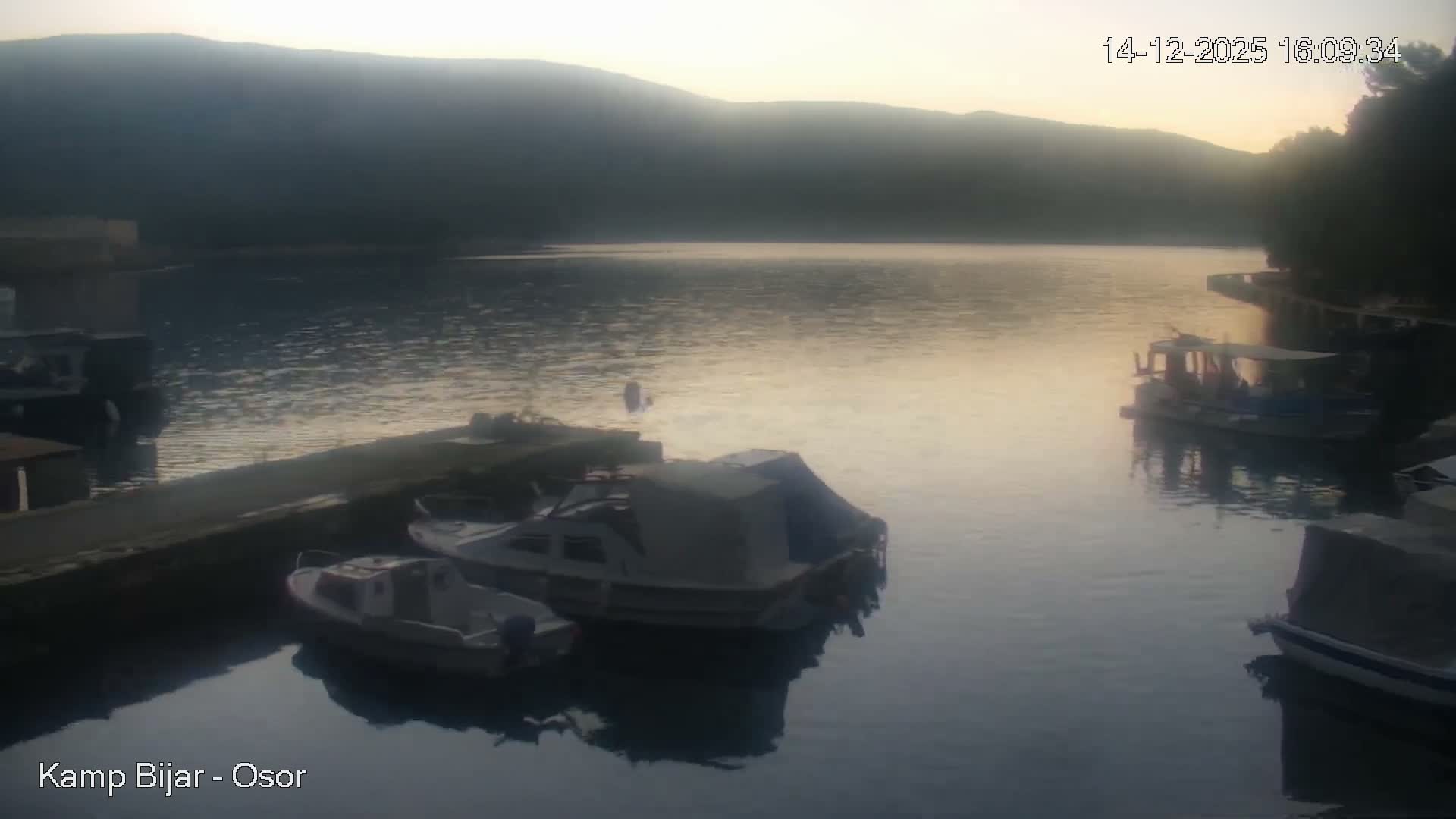 Several boats are moored in a tranquil bay with stone docks and lush hills, under an overcast and rainy sky.