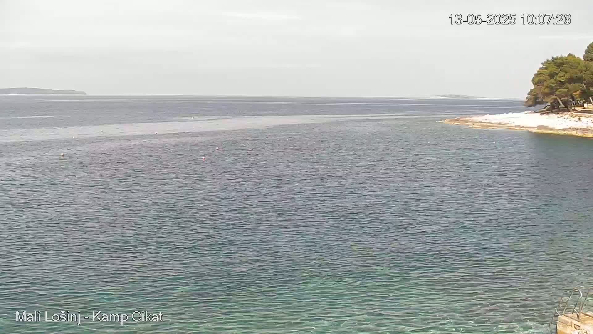 A calm, overcast day shows a tranquil sea with a small, tree-lined shoreline in the foreground and a distant island on the horizon.