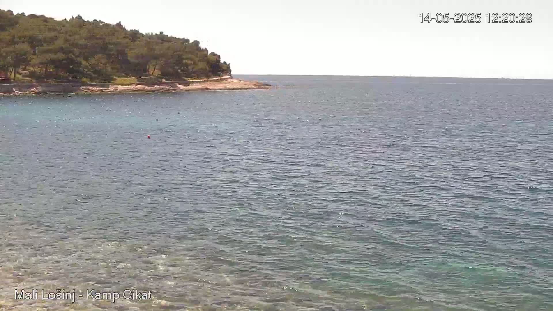 A calm, clear body of water laps against a rocky shoreline where a small, tree-covered hill sits.