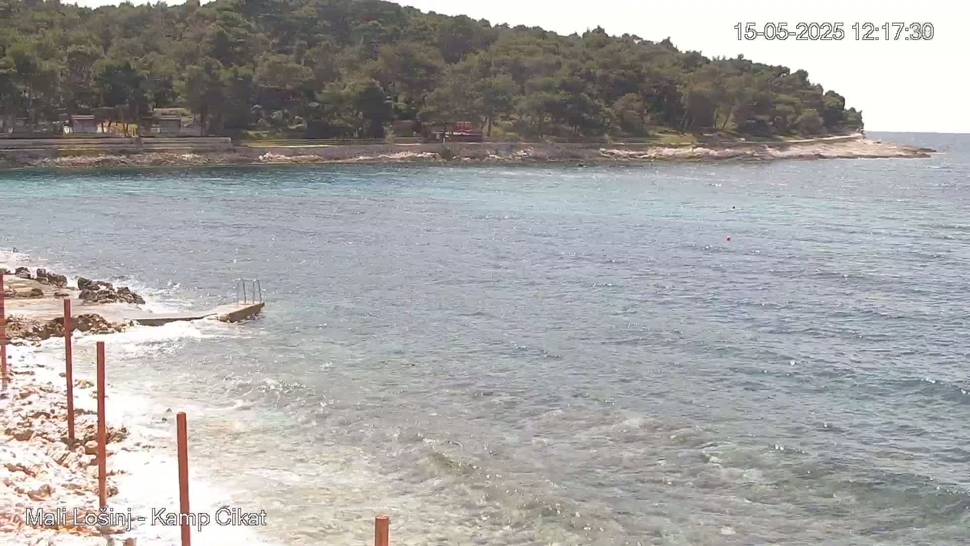 A rocky shoreline with a small concrete ramp leading into calm, shallow, blue-green water, backed by a green, tree-covered hill on a sunny day.