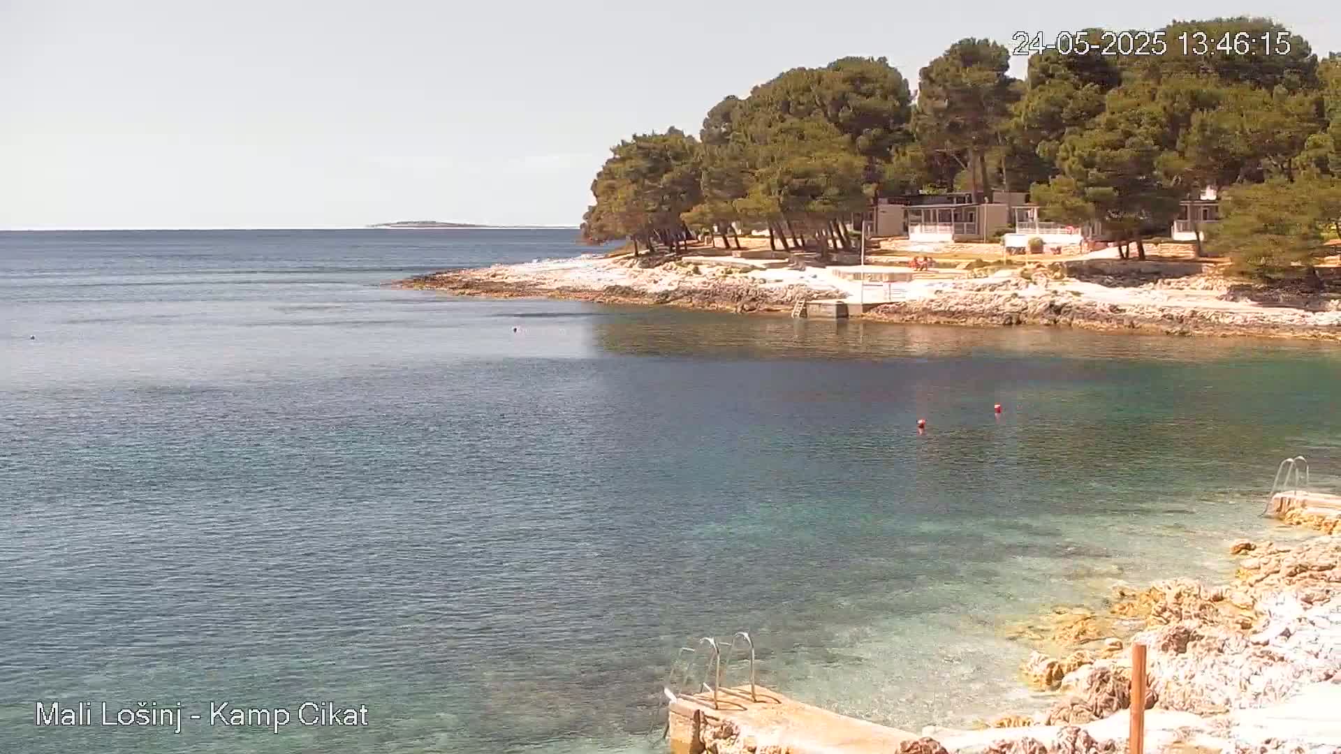 A tranquil bay with clear, shallow water laps against a rocky shore lined with pine trees and small buildings under a sunny sky.