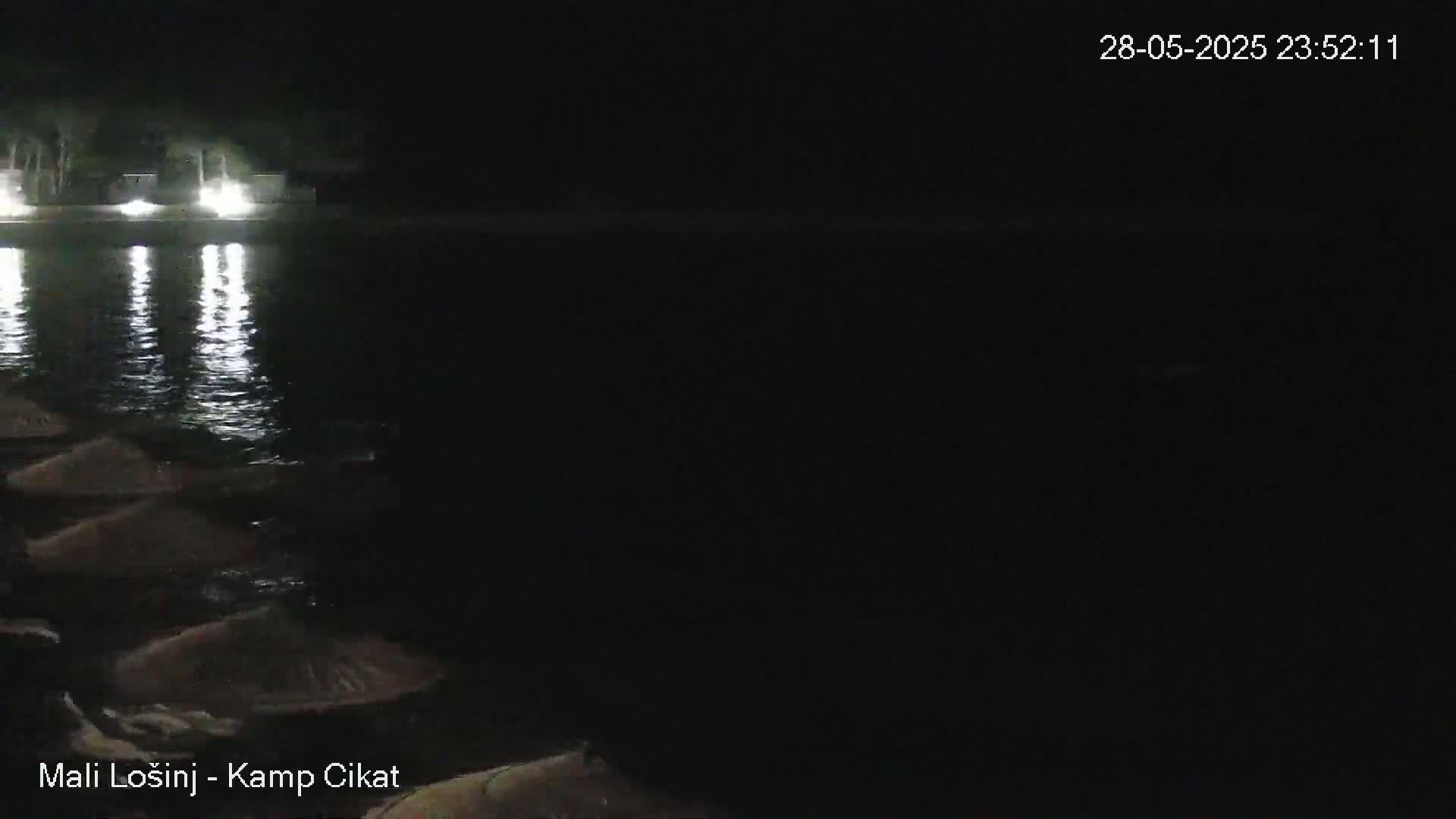 At night, a dark body of water reflects lights from a shoreline with several dark objects, possibly umbrellas, arranged along the water's edge.