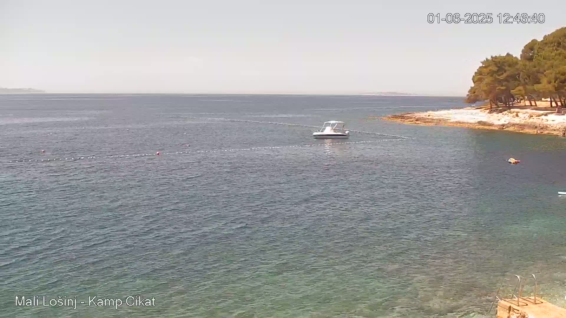 A small motorboat is anchored in calm, clear water near a rocky shoreline with trees in the background under a bright, sunny sky.