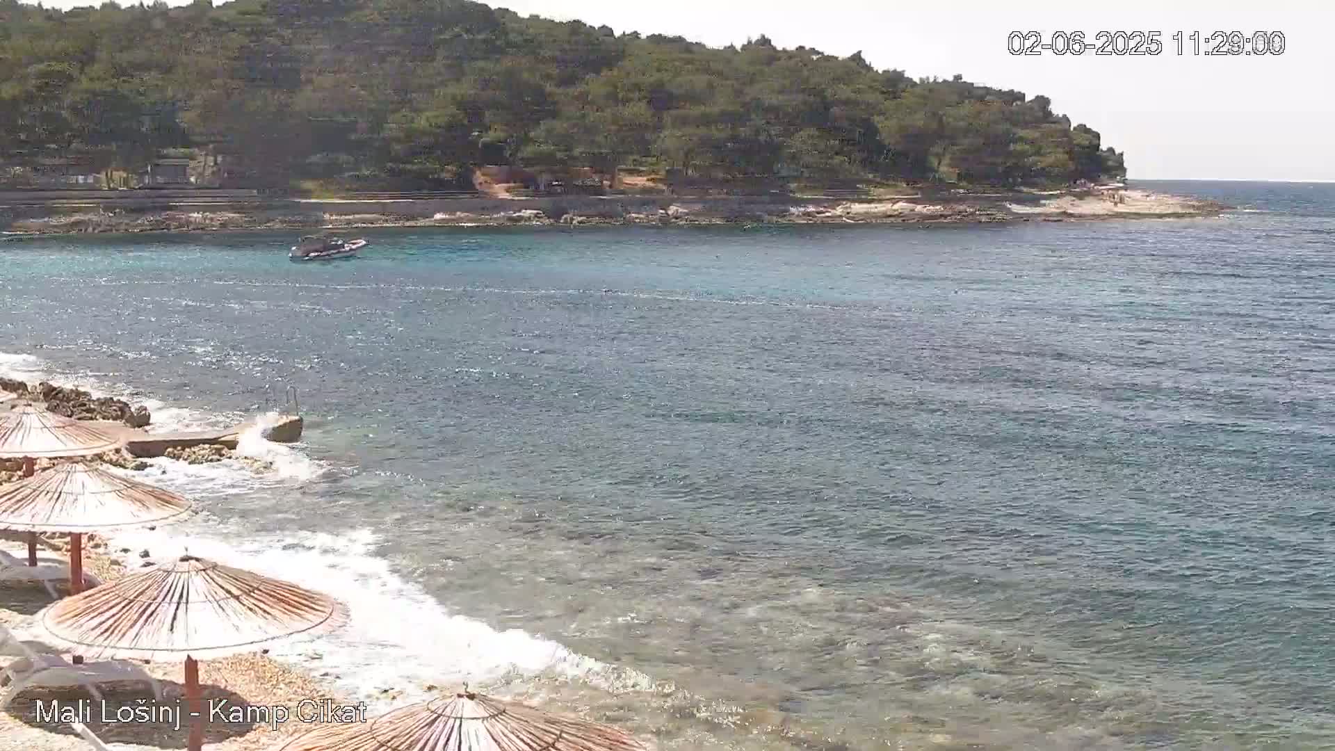 A beach with several straw umbrellas is shown, with calm waves lapping the shore, a small boat on the water, and a tree-covered hill in the background under a sunny sky.