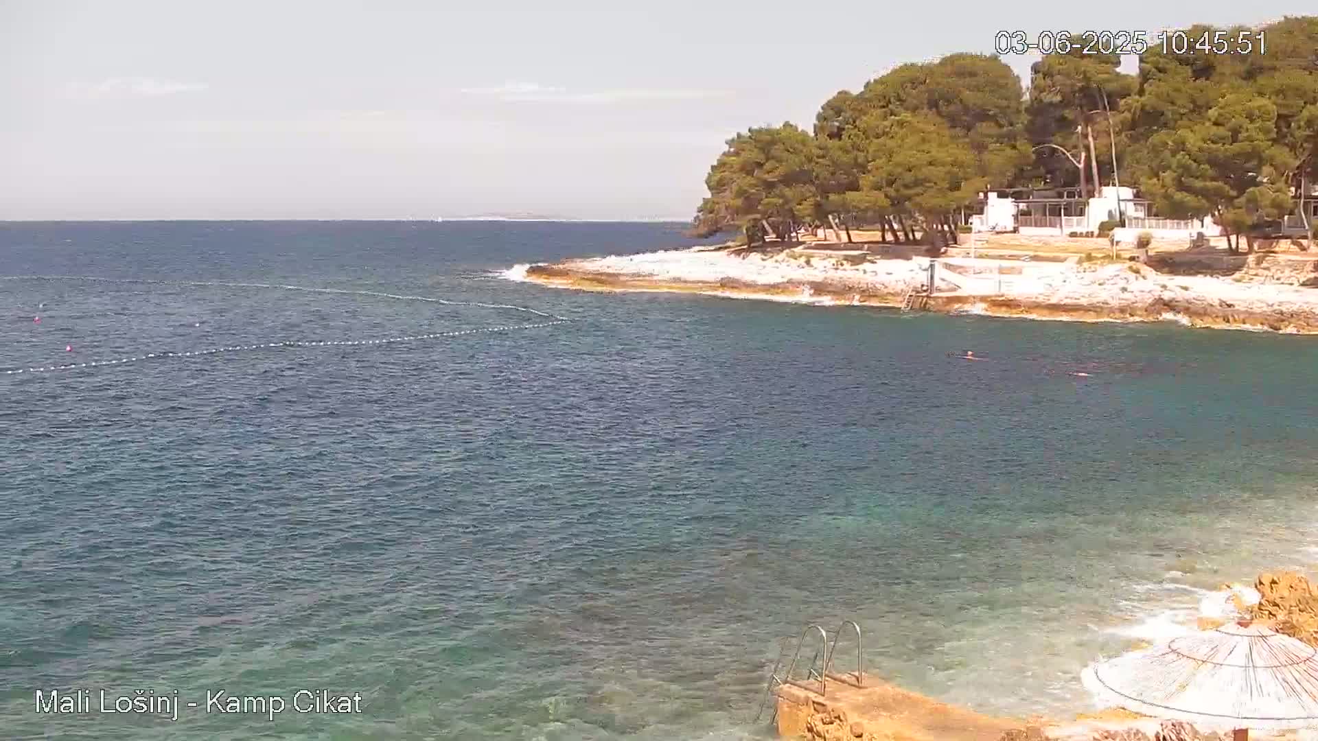 A calm, clear ocean bay with a rocky shore lined with trees and a few buildings under a sunny sky.