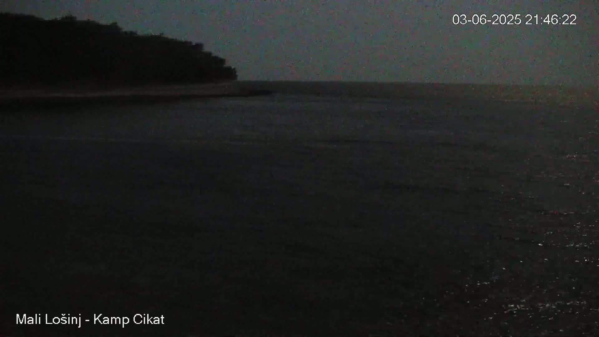 A dark, nighttime view shows a body of water with a shoreline featuring a silhouetted landmass under a dusky sky.
