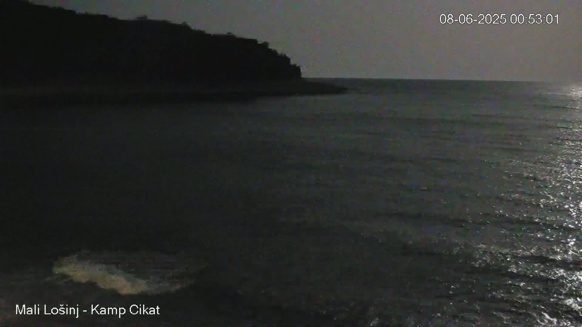 A dark, nighttime coastal scene shows a calm sea under a dim sky, with a silhouetted shoreline in the background.