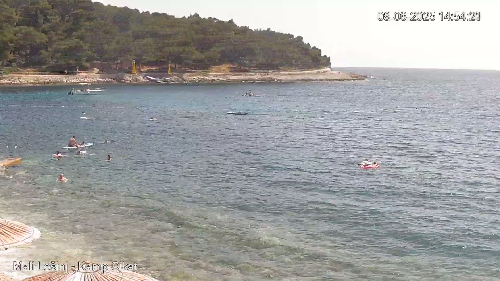 A calm, clear day at a beach shows people swimming and paddleboarding in shallow, clear water near a shoreline with green trees and hills.