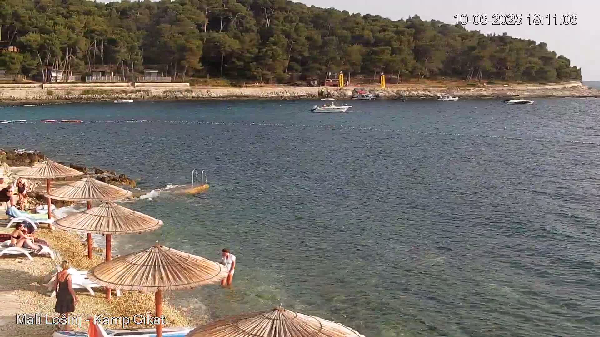 A rocky beach with several straw umbrellas and people relaxing, next to calm water with boats and a wooded hillside in the background.
