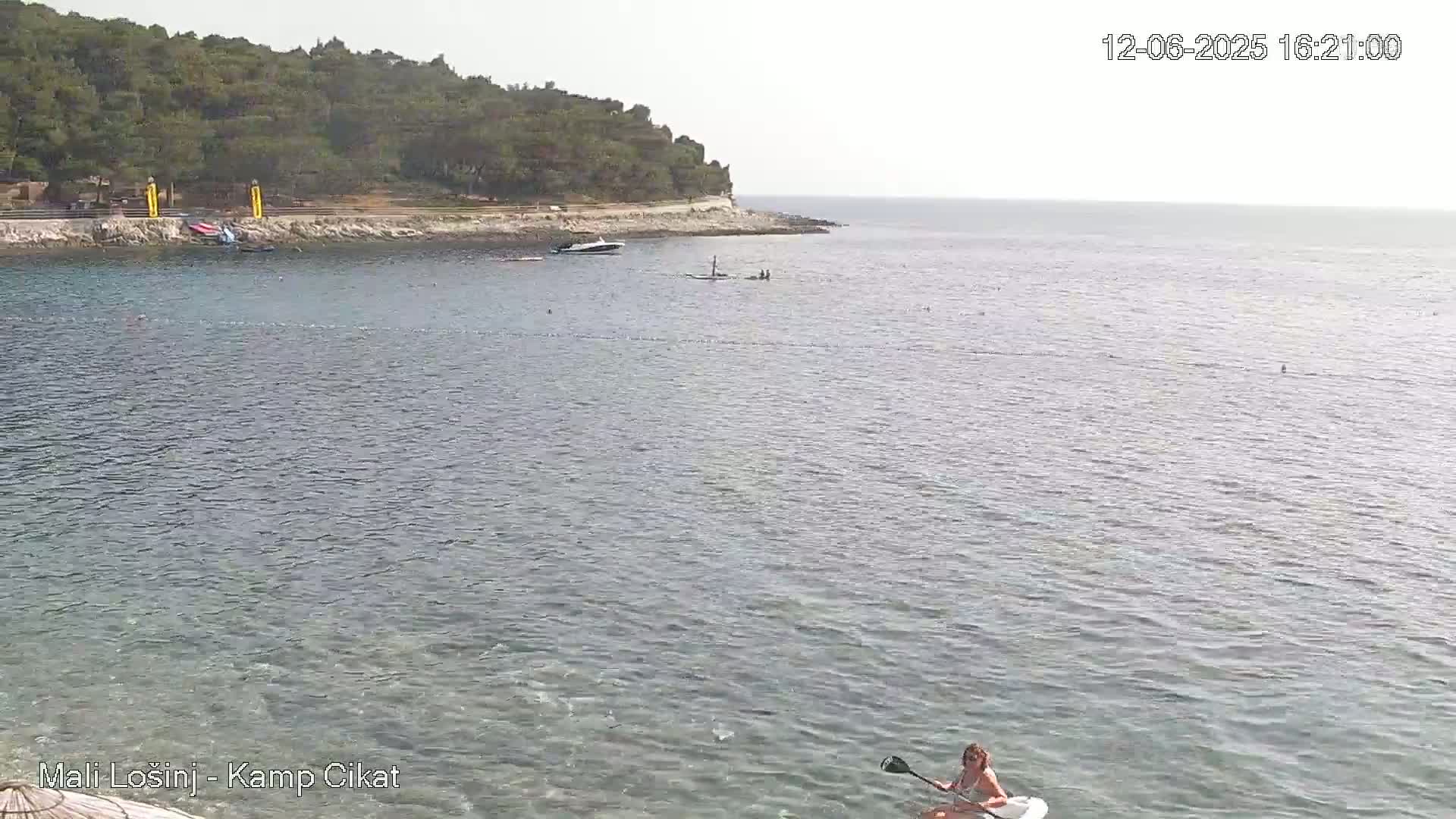 A woman paddleboards in calm, clear water near a rocky shoreline with green trees under a bright, sunny sky.