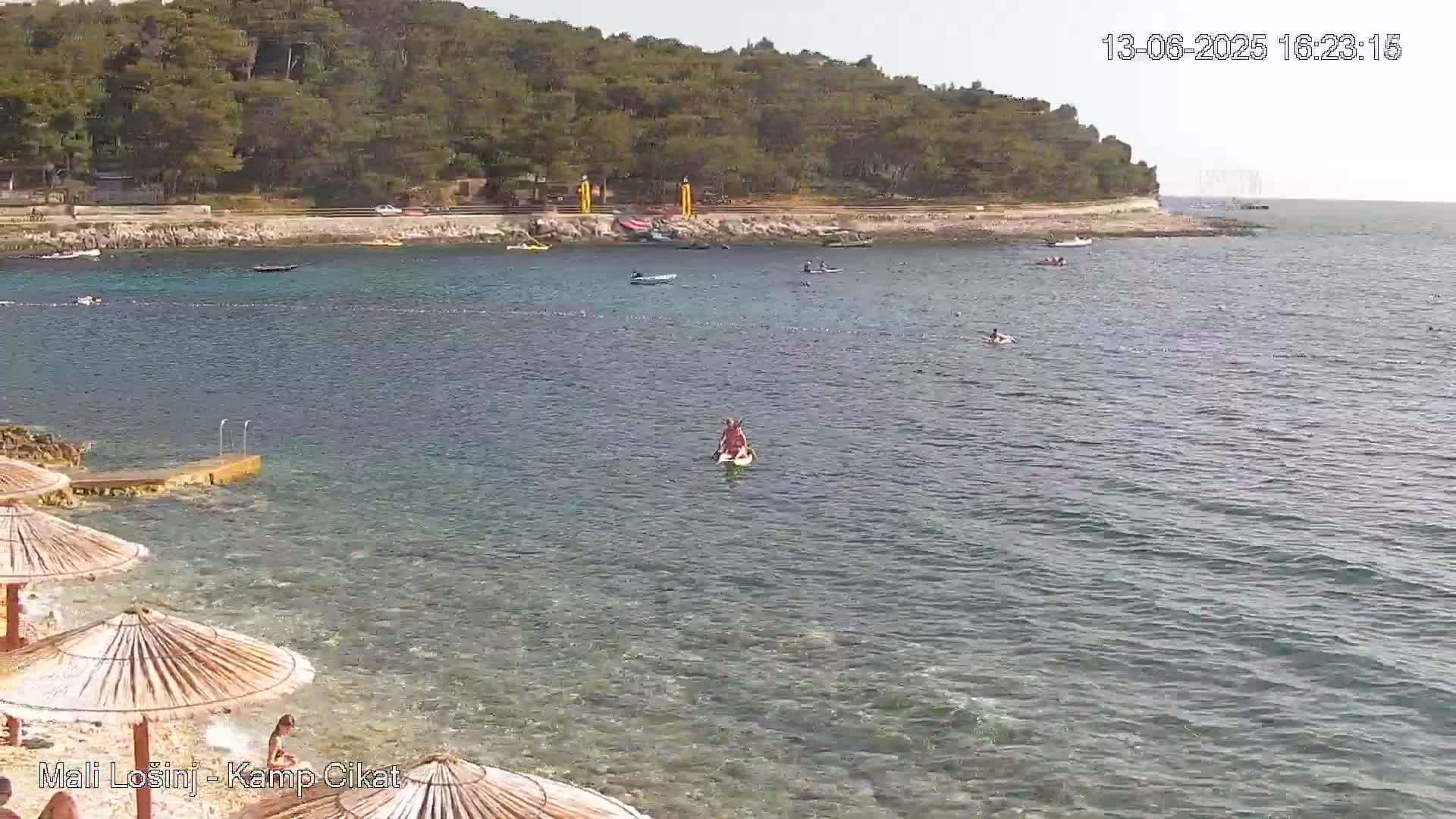 A sunny day at a calm beach shows several small boats and people swimming or paddleboarding in clear, shallow water near a rocky shore lined with trees and umbrellas.