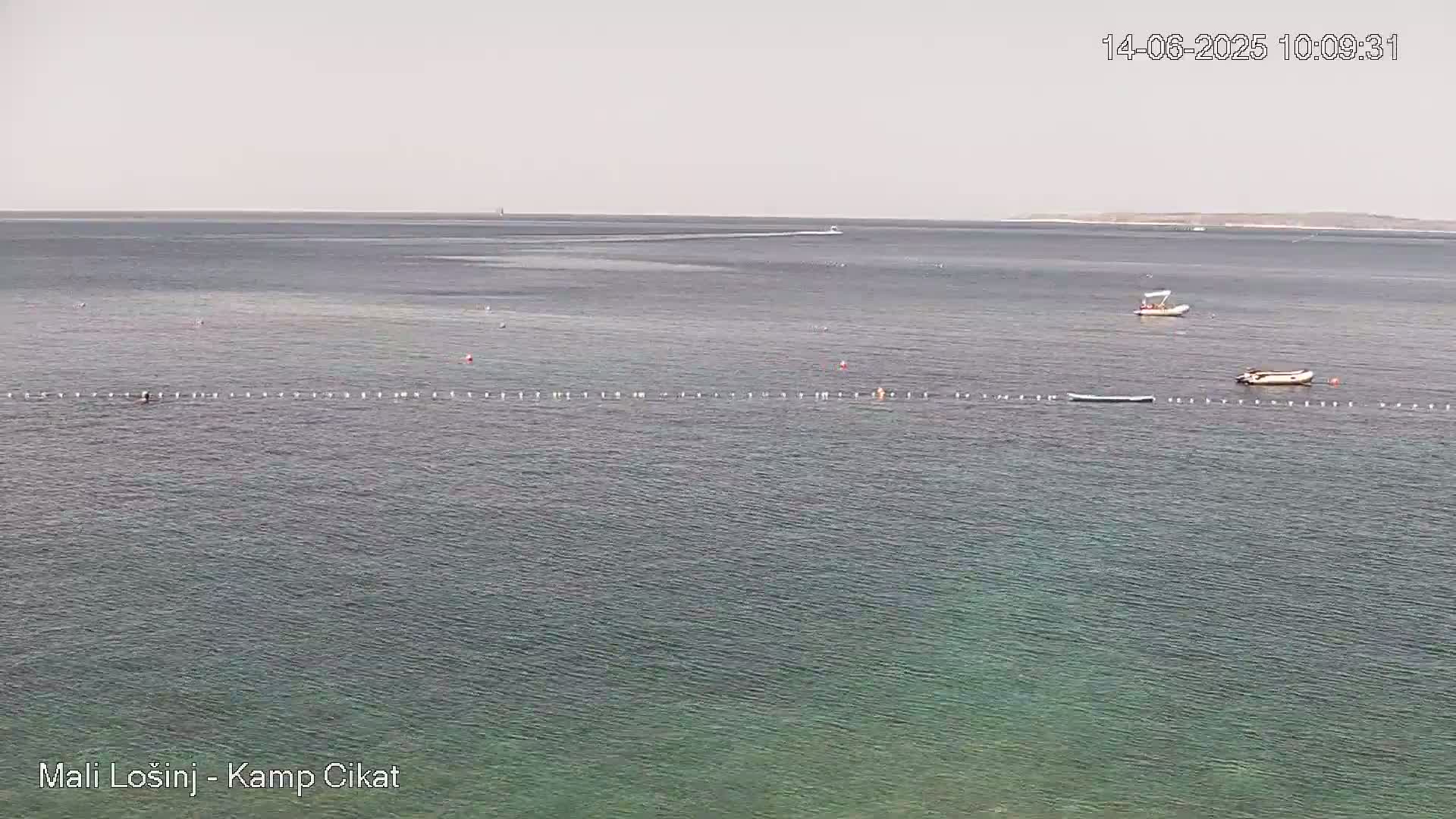 A calm, light-grey sea with a few small boats and a long line of buoys under a hazy sky.
