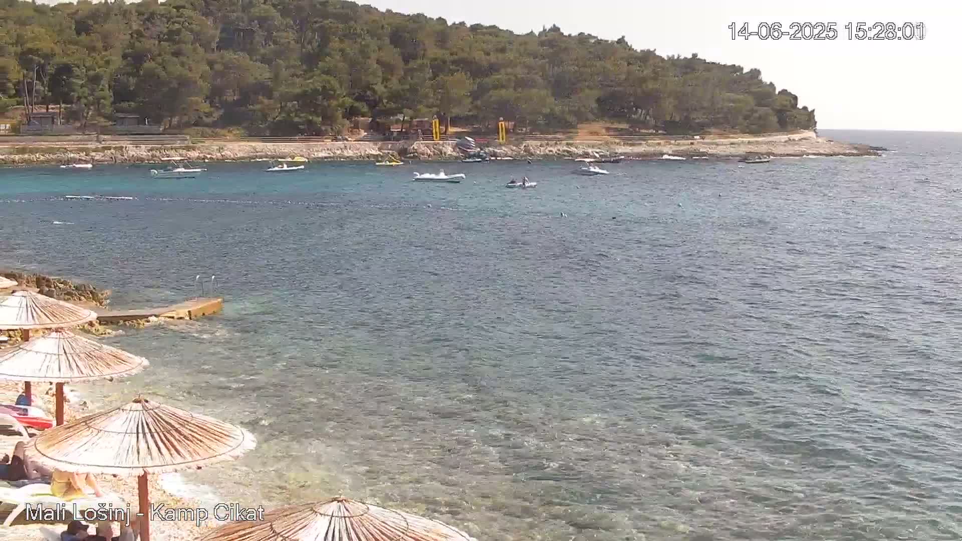 A calm, sunny day at a beach with several small boats in the water, a rocky shore with straw umbrellas, and a green hill in the background.