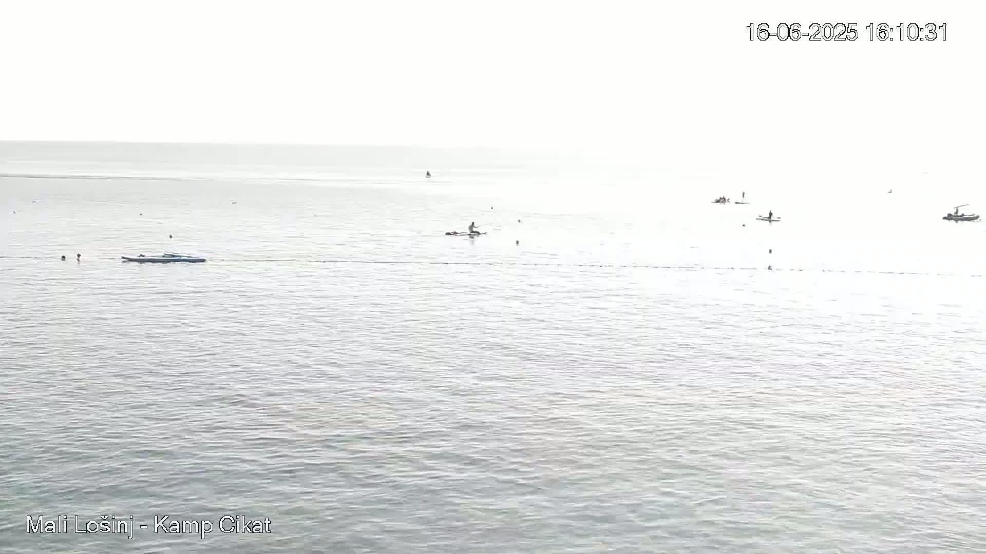 A calm, light-colored body of water is sparsely populated with several small watercraft and swimmers on a hazy day.