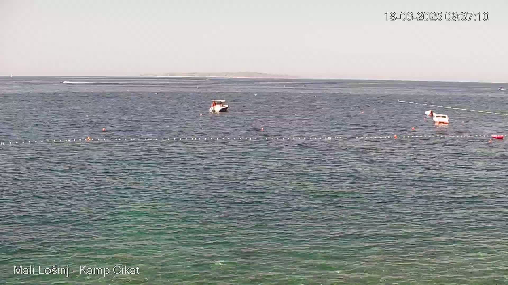 Calm, clear water with two small boats and a line of buoys in the foreground, and a distant landmass under a pale sky.