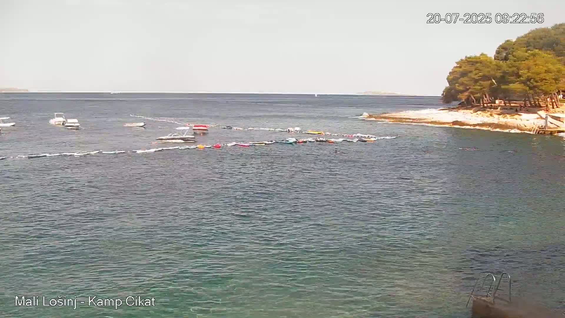 Calm, clear water with several small boats and inflatable floats near a rocky shoreline with trees under a mostly sunny sky.