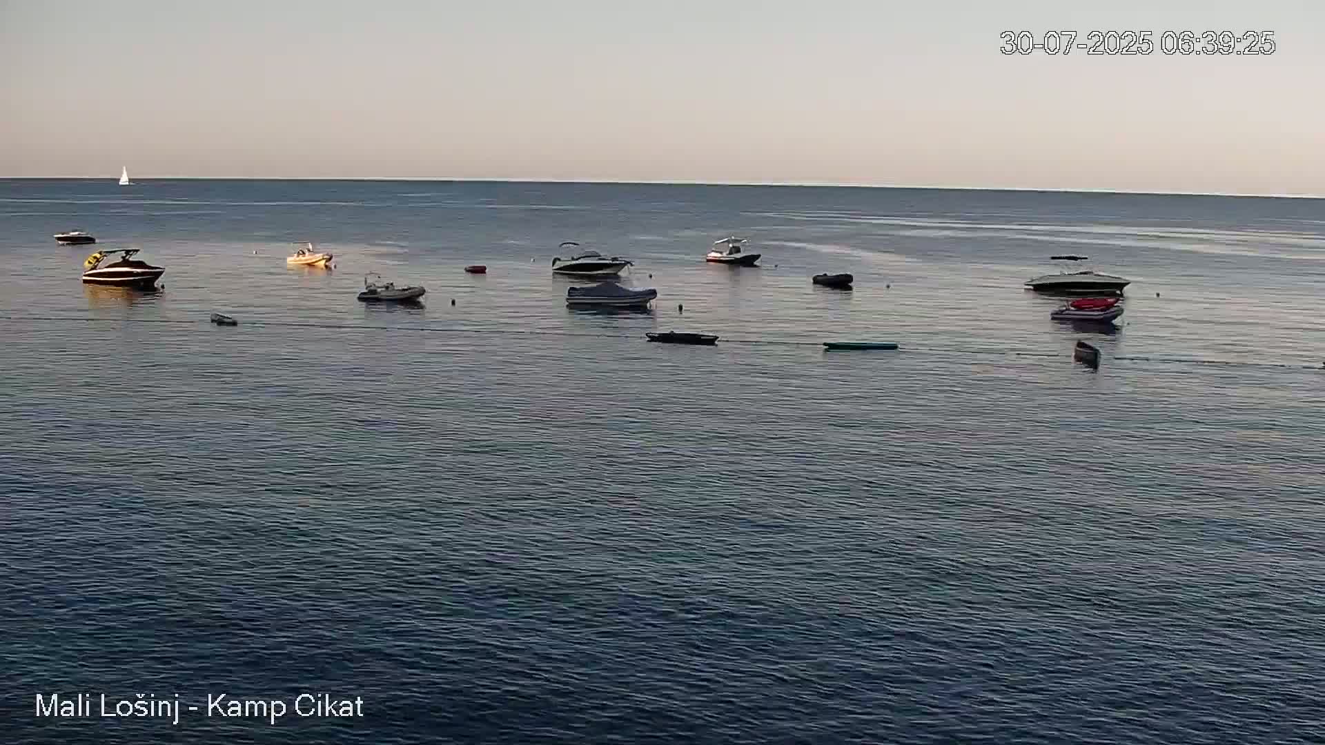 Calm, clear water with several small boats and inflatable floats near a rocky shoreline with trees under a mostly sunny sky.