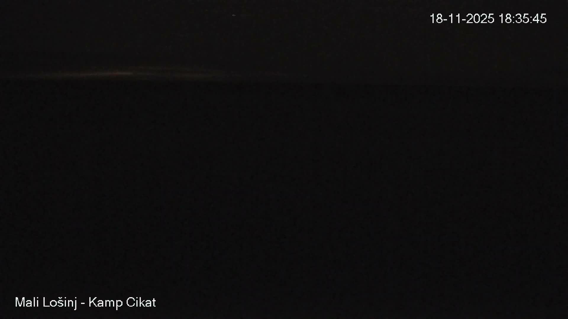 A calm, sunny day shows a clear bay with several boats and people swimming, near a shoreline lined with green trees.