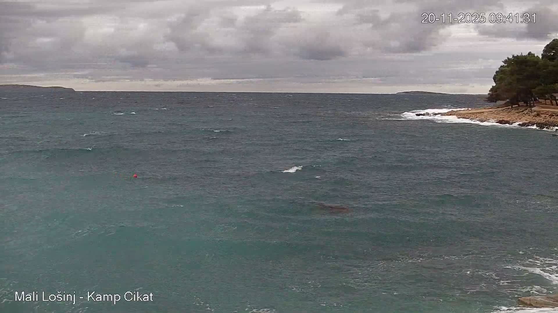 Choppy teal-blue sea water with a red buoy, under an overcast sky, meets a rocky, tree-lined coast with breaking waves and a distant island visible on the horizon.
