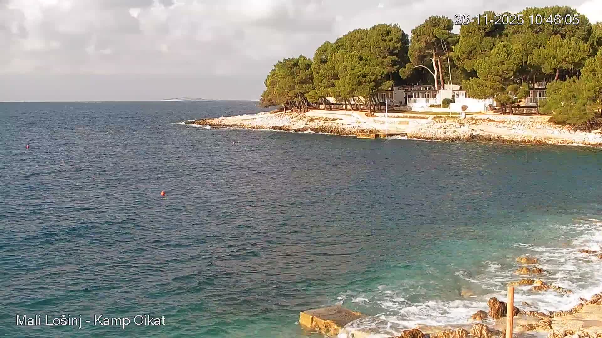 A rocky coastline densely forested with green pine trees and scattered white buildings borders a clear blue-green sea with gentle waves, under a partly cloudy sky, with two orange buoys visible in the water and a distant landmass on the horizon.