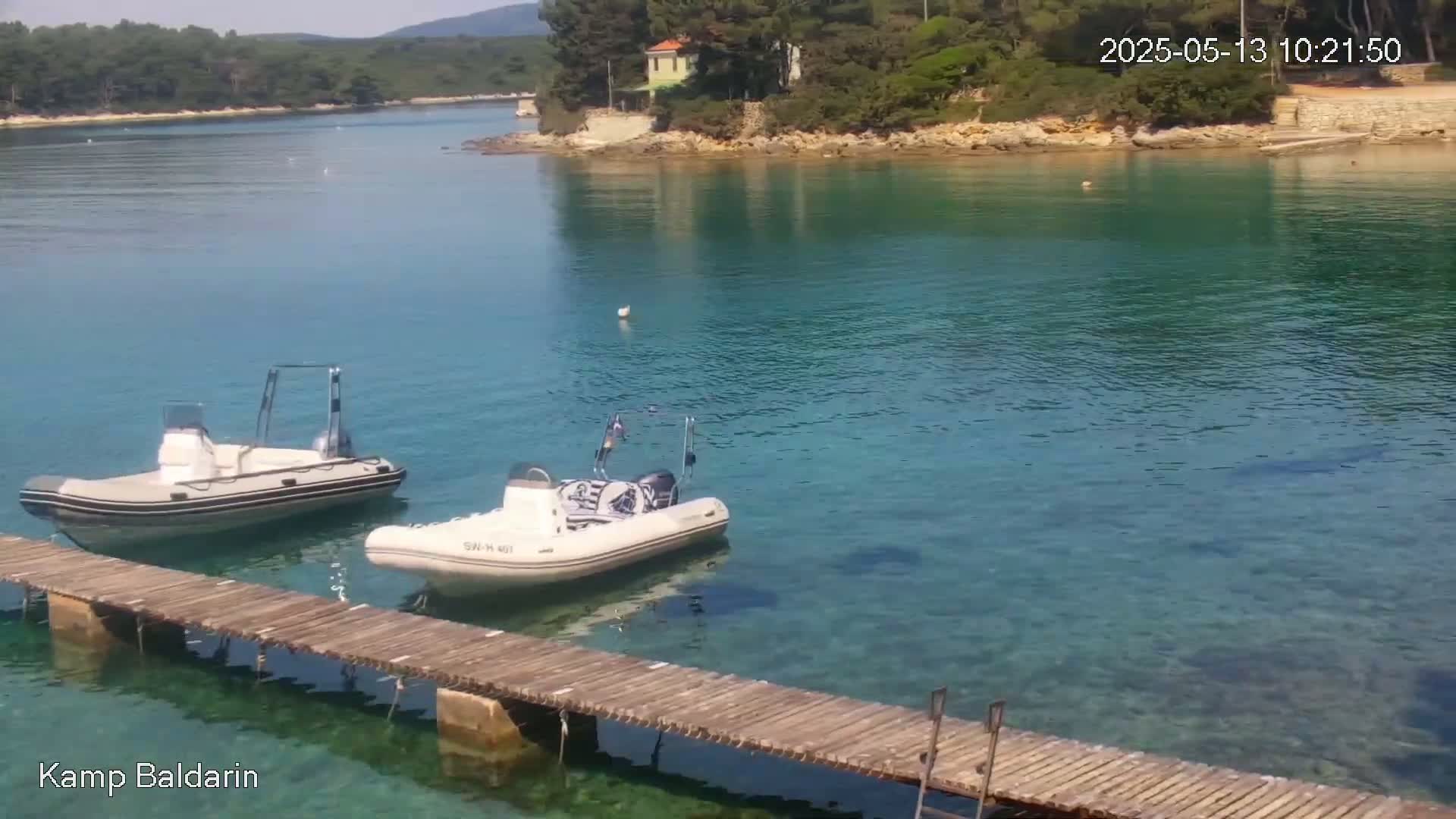 Two inflatable motorboats are moored at a wooden dock in a calm, clear turquoise bay, with green hills and trees in the background.