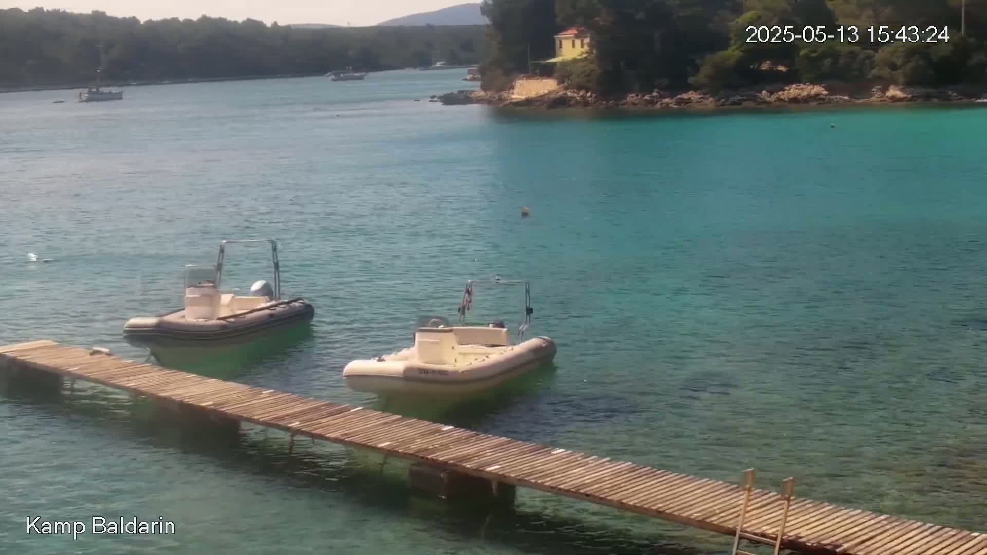 Two inflatable boats are moored at a wooden dock in calm, clear turquoise water, with a small yellow building visible on the shoreline in the background.