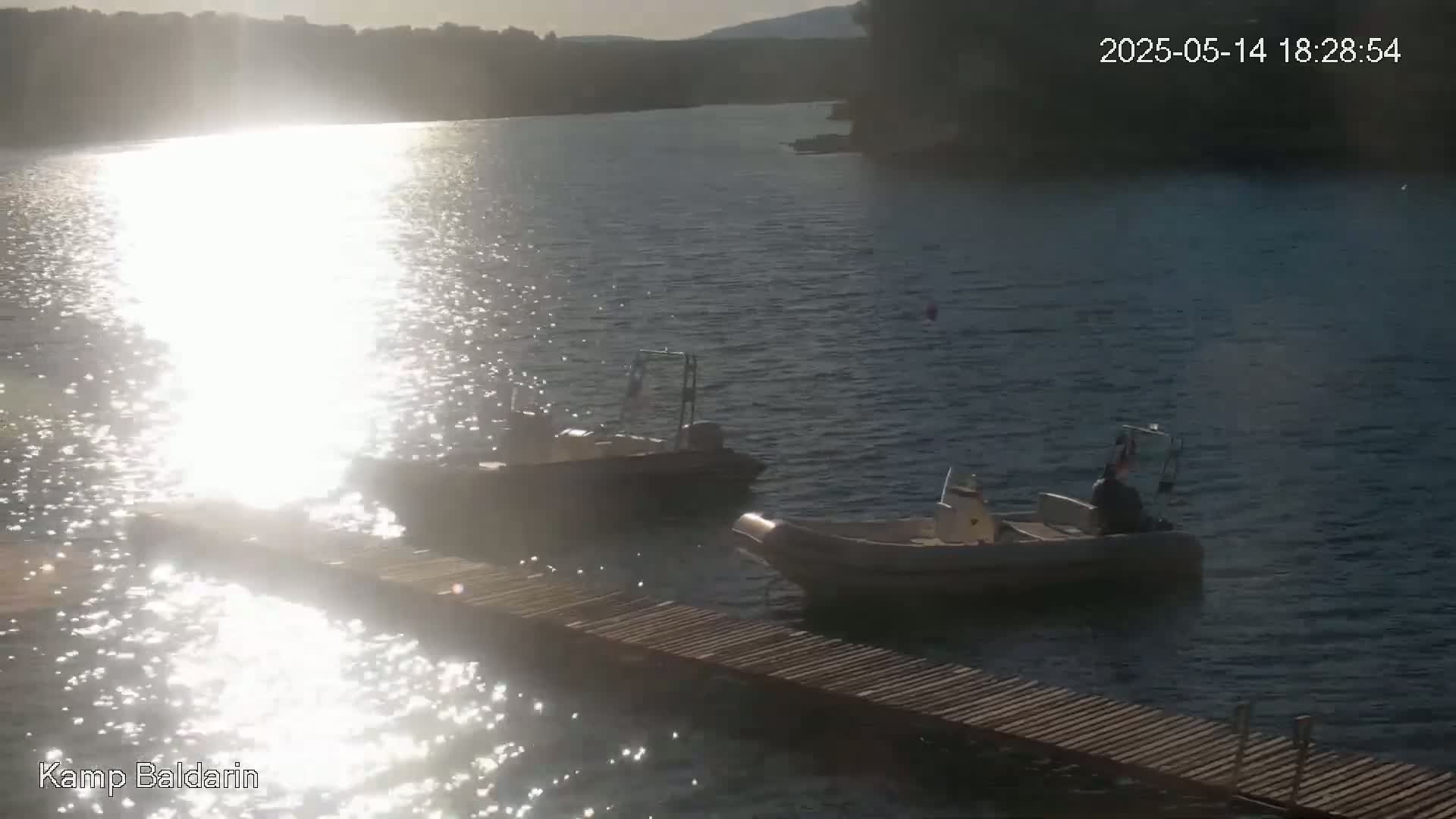 Two small motorboats are moored near a wooden dock on a calm body of water, with sunlight reflecting brightly on the surface.